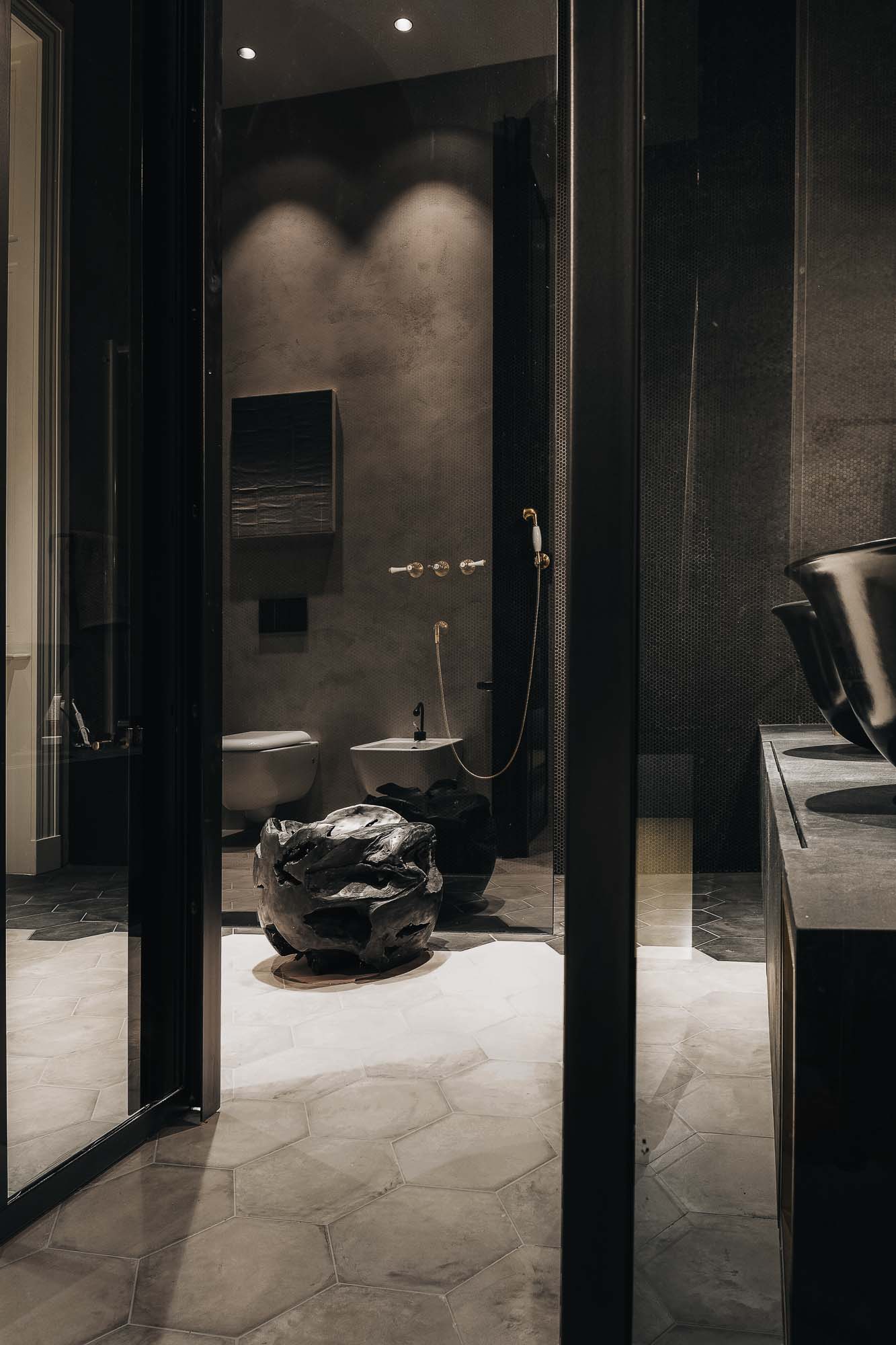 Černá koupelna bytu Pařížská – pohled přes prosklené příčky, hexagonální dlaždice, skulpturální keramický objekt na podlaze, bidet a toaleta v pozadí, černé mísovité umyvadlo vpravo