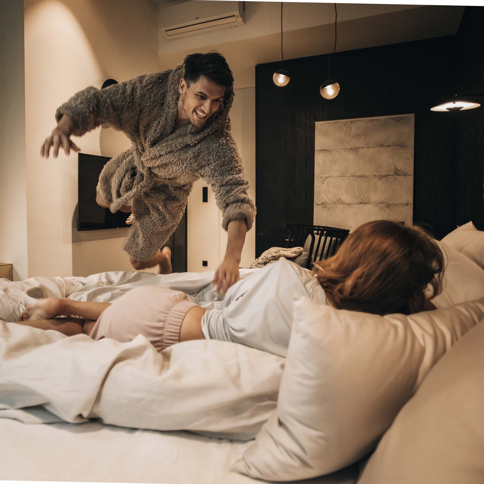 Lifestyle fotografie v ložnici bytu Pařížská – muž v županu skáče na postel k ležící ženě, tmavé stěny s dřevěným obkladem v pozadí, visící kulové světla