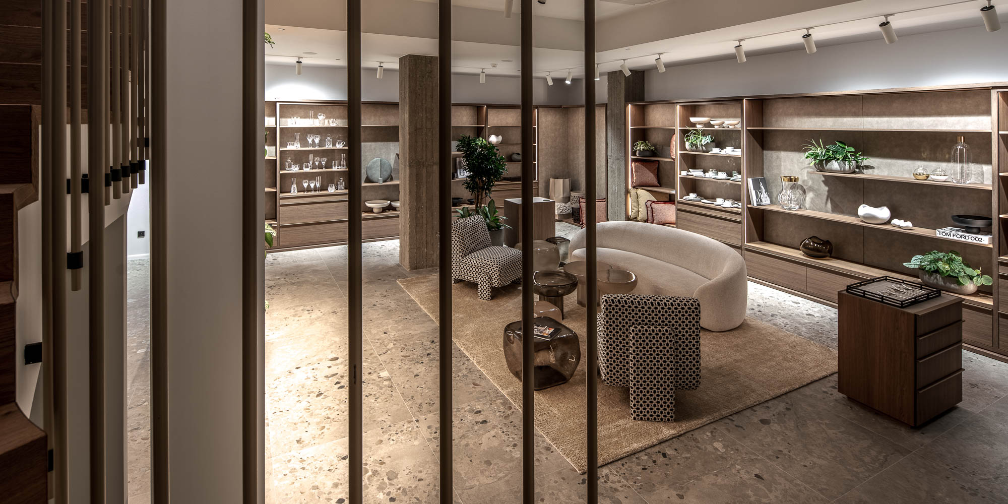Pohled ze schodiště skrze tyčkové zábradlí do showroomu LA TABLE Pařížská s výstavními policemi, sezónní zónou a produkty – OOOOX design