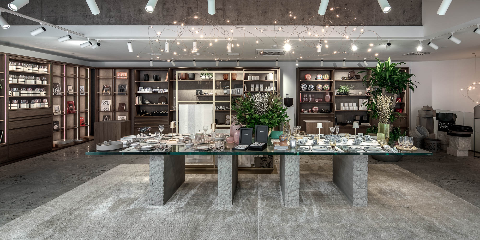 Panoramatický pohled na showroom LA TABLE Pařížská – centrální skleněný stůl plně prostřený, výstavní police s produkty a rostlinami po celém obvodu – OOOOX