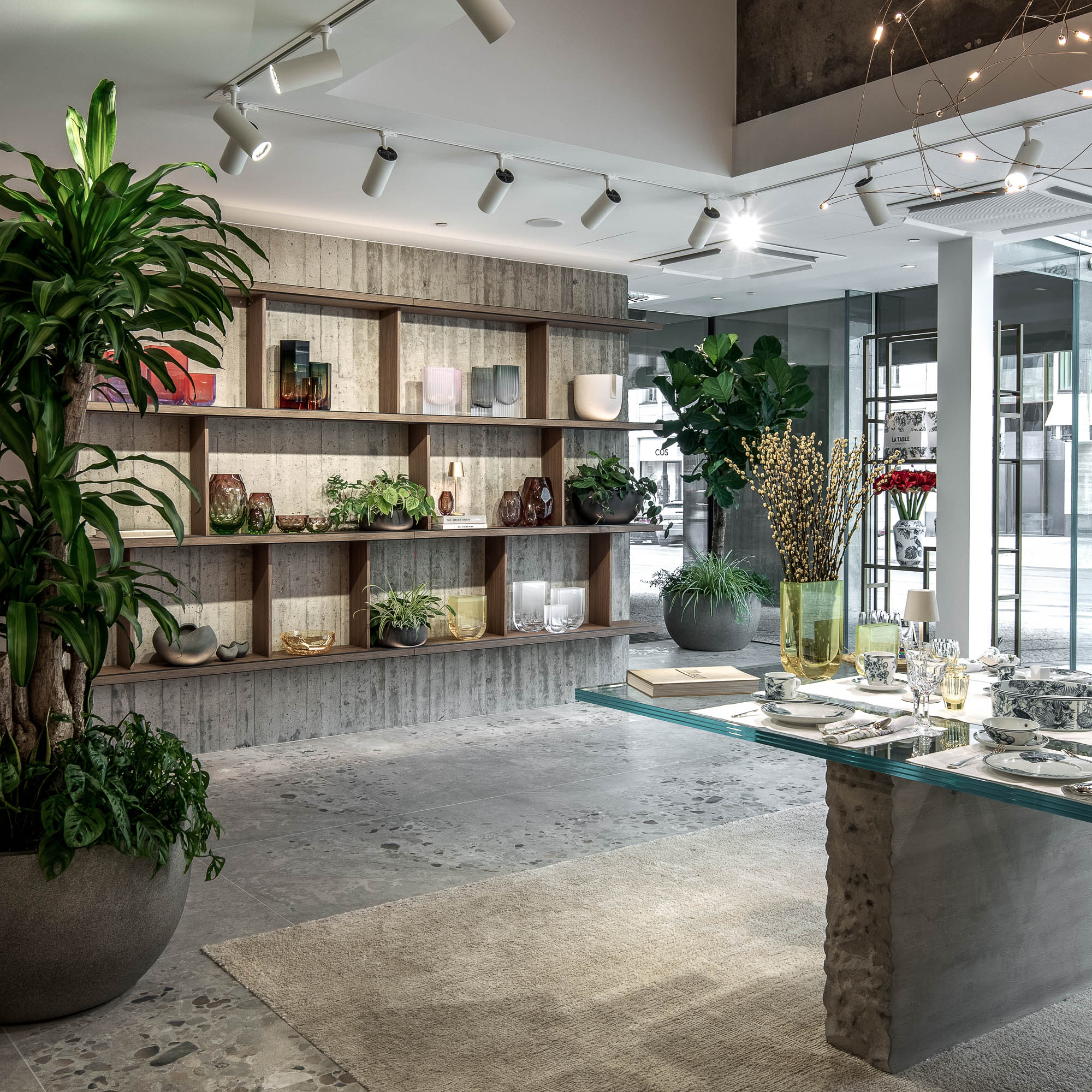 Pohled na showroom LA TABLE Pařížská se skleněným stolem, výstavními policemi s produkty, prosklenou fasádou a velkými pokojovými rostlinami v popředí – OOOOX