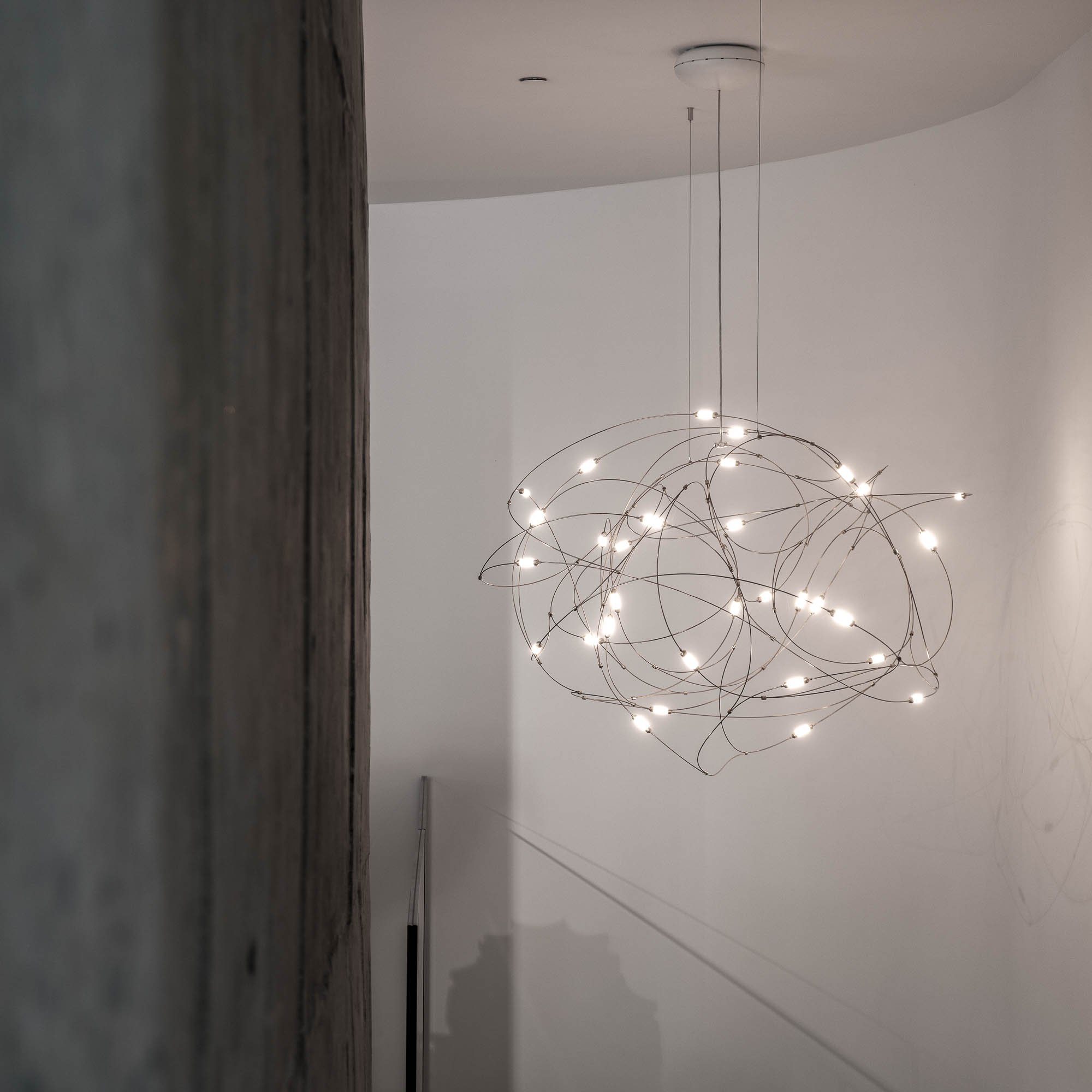 Detail závěsného svítidla Moooi Flock of Light v schodišťovém prostoru butiku LA TABLE Pařížská – přiznané betonové pilíře a zakřivená bílá stěna – OOOOX