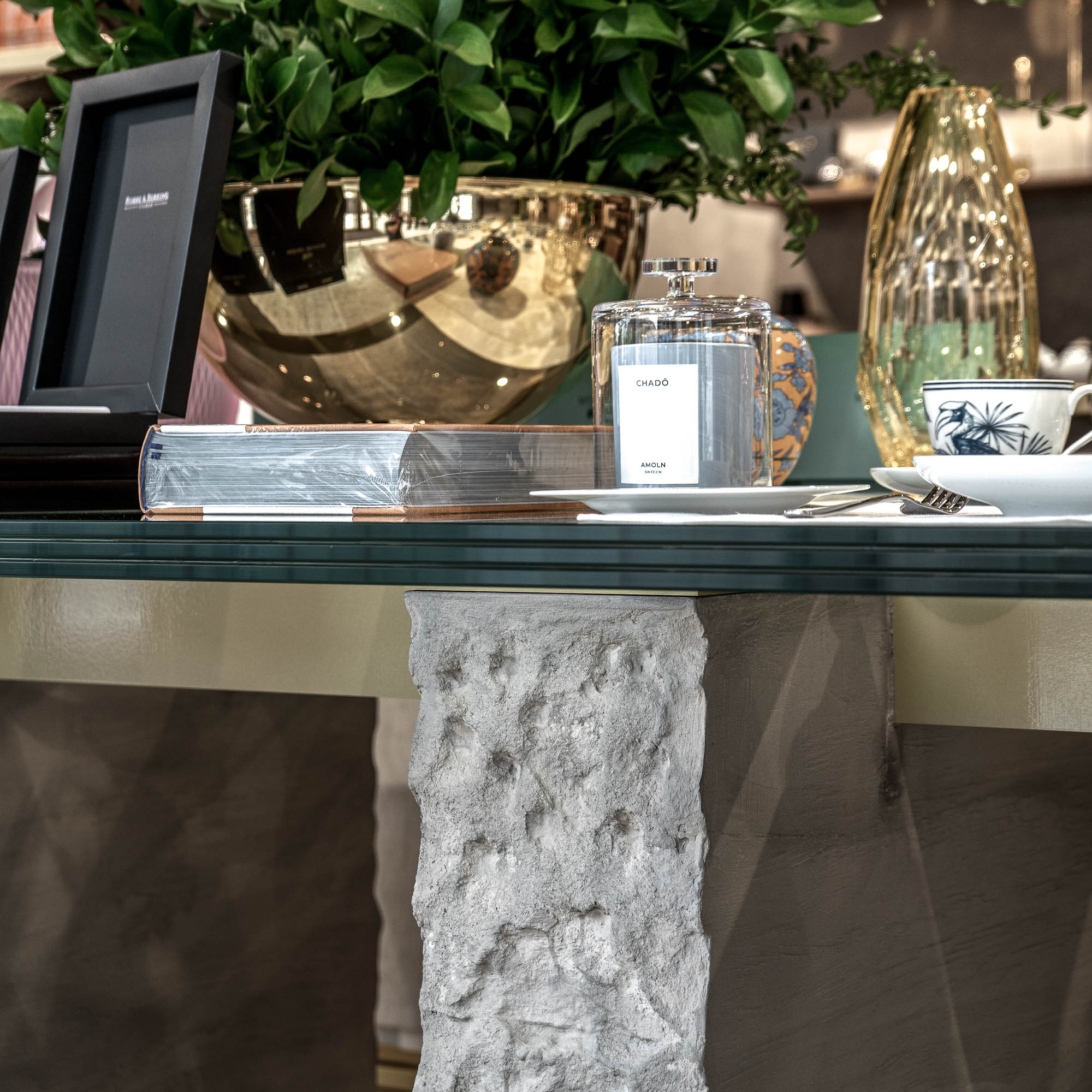 Detail skleněného stolu showroomu LA TABLE Pařížská s luxusními produkty – parfém Chado, zlatá miska, skleněná váza a kávová souprava na trojité skleněné desce – OOOOX