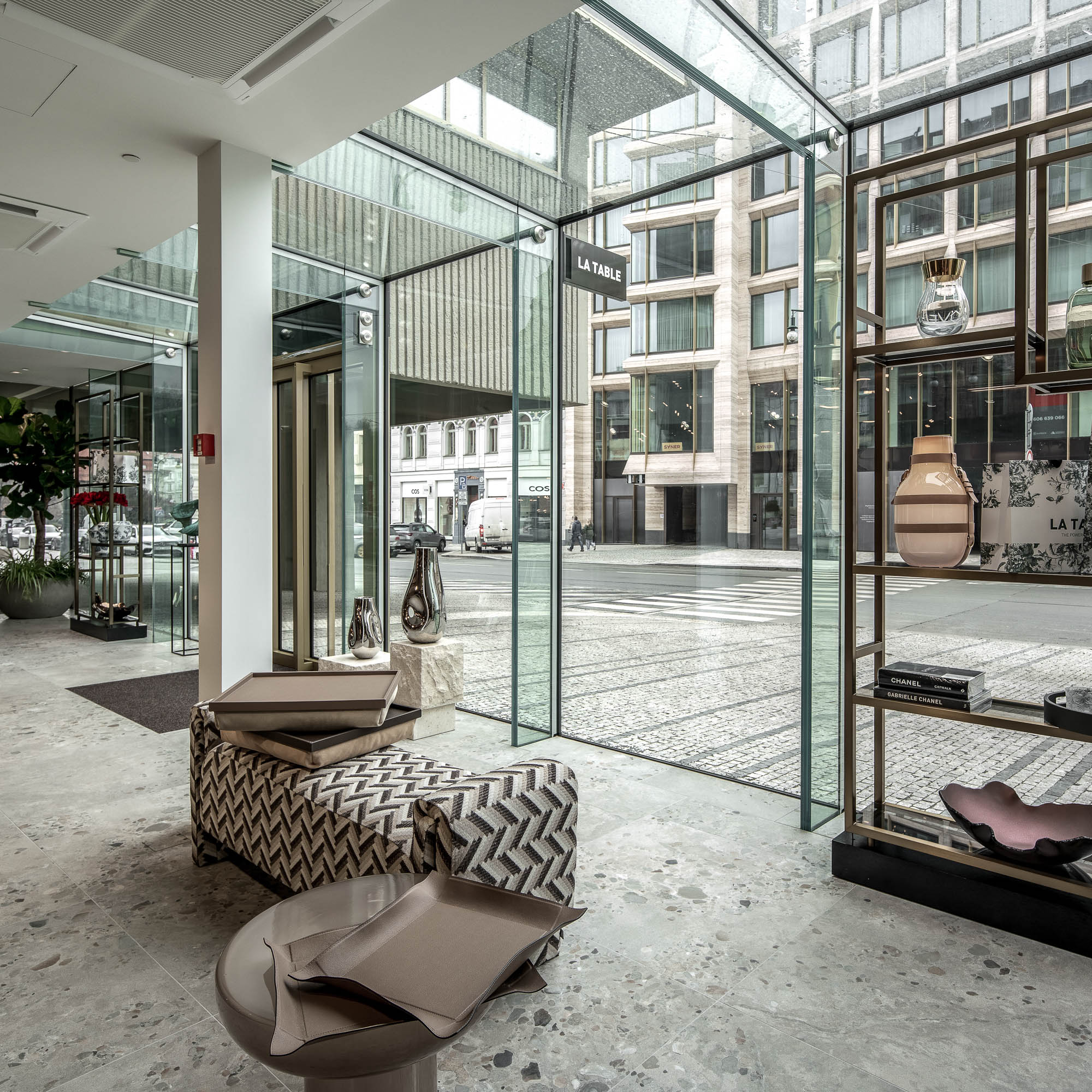 Vstupní lobby butiku LA TABLE Pařížská v hotelu Fairmont – skleněný box vstupu, vzorovaný taburet, výhled na ulici Pařížská a výstavní police – OOOOX design