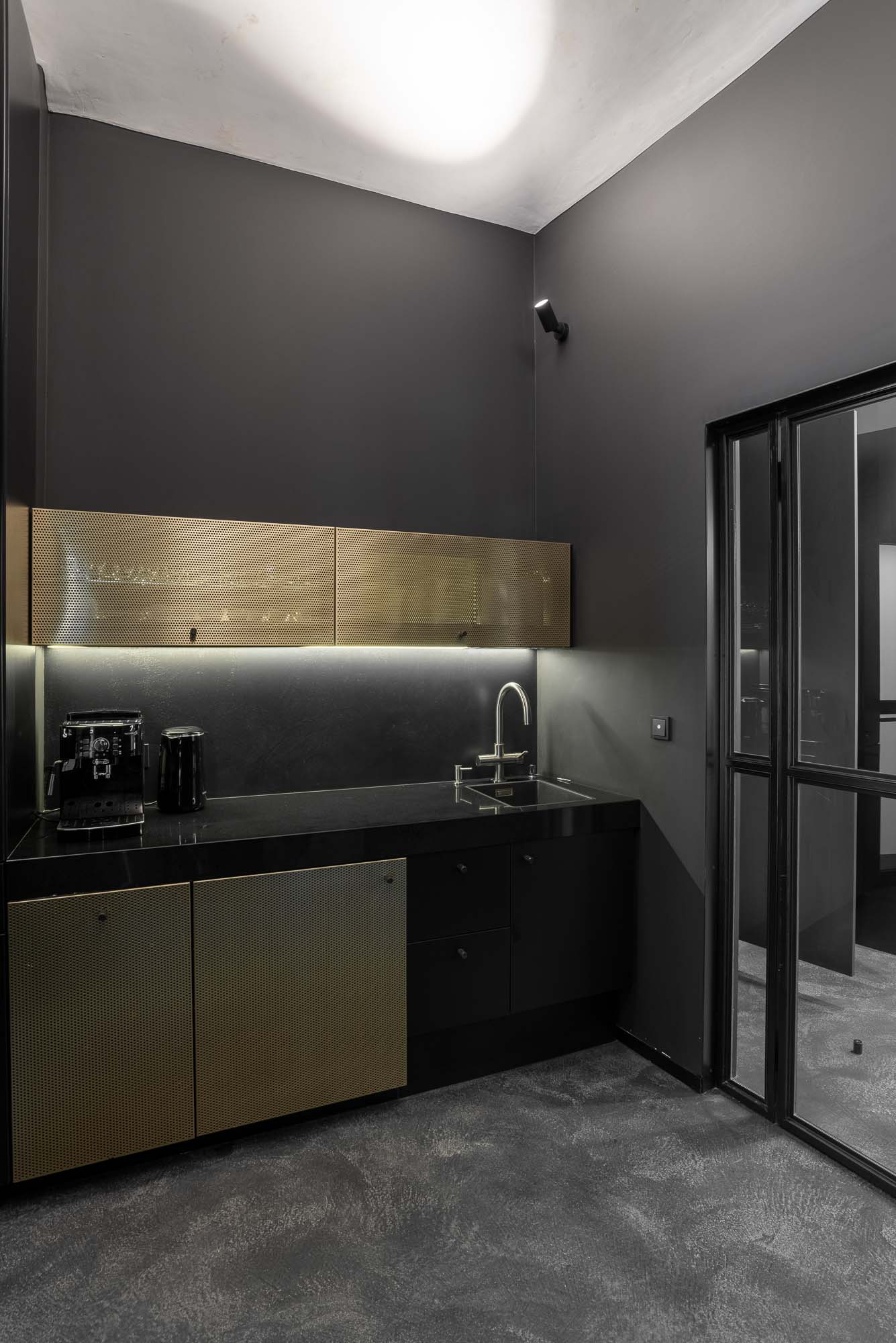 Černá designová kuchyňka s mosaznými perforovanými dvířky, industriálními materiály a nábytkem vyrobeným na míru v moderním kancelářském interiéru. Design by OOOOX