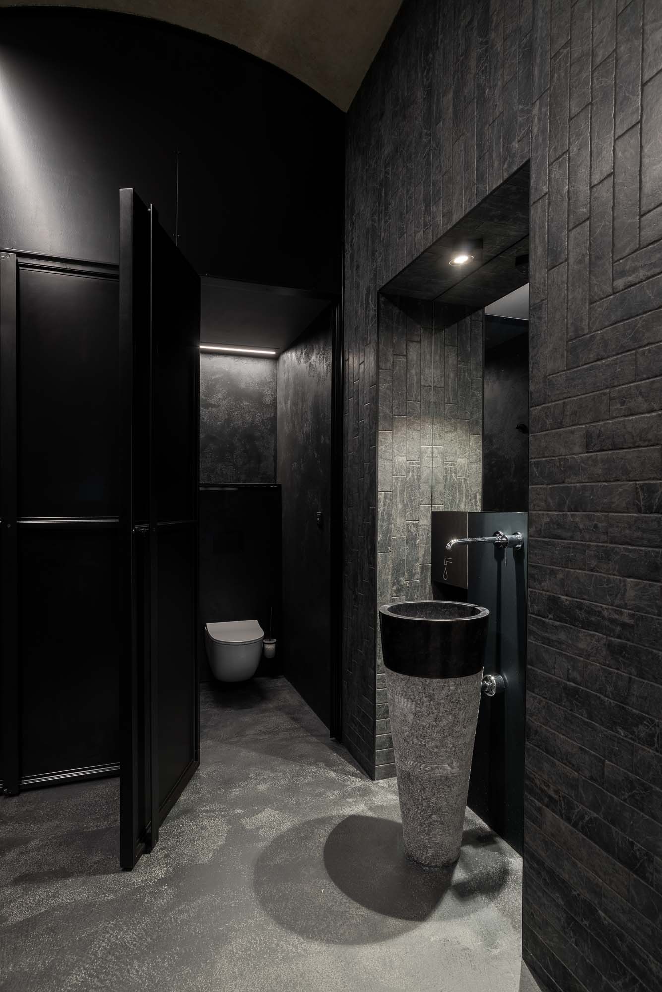 Detail kamenného stojícího umyvadla v černém industriálním interiéru, designové baterie, velkého nástěnného zrcadla s decentním osvětlením a keramického obkladu imitujícího přírodní kámen. Design by OOOOX