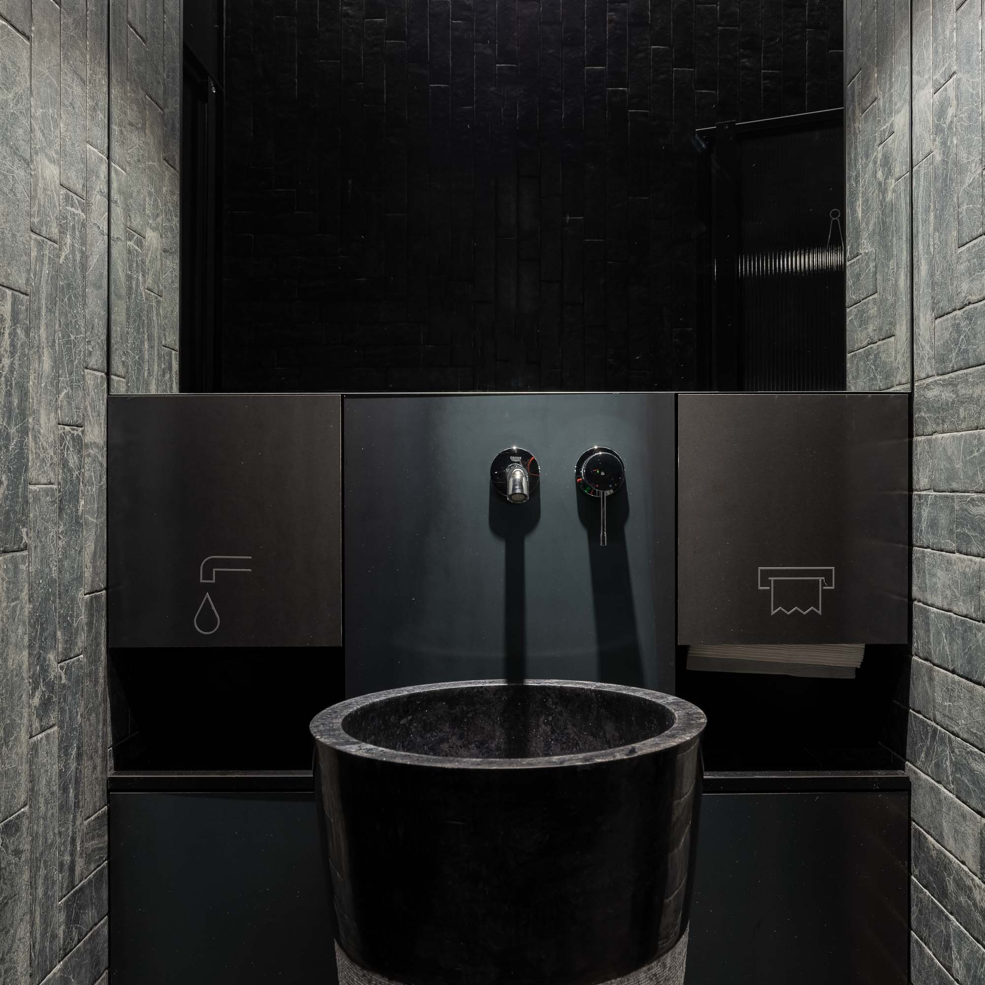 Detail kamenného stojícího umyvadla v černém industriálním interiéru, designové baterie, velkého nástěnného zrcadla s decentním osvětlením a keramického obkladu imitujícího přírodní kámen. Design by OOOOX