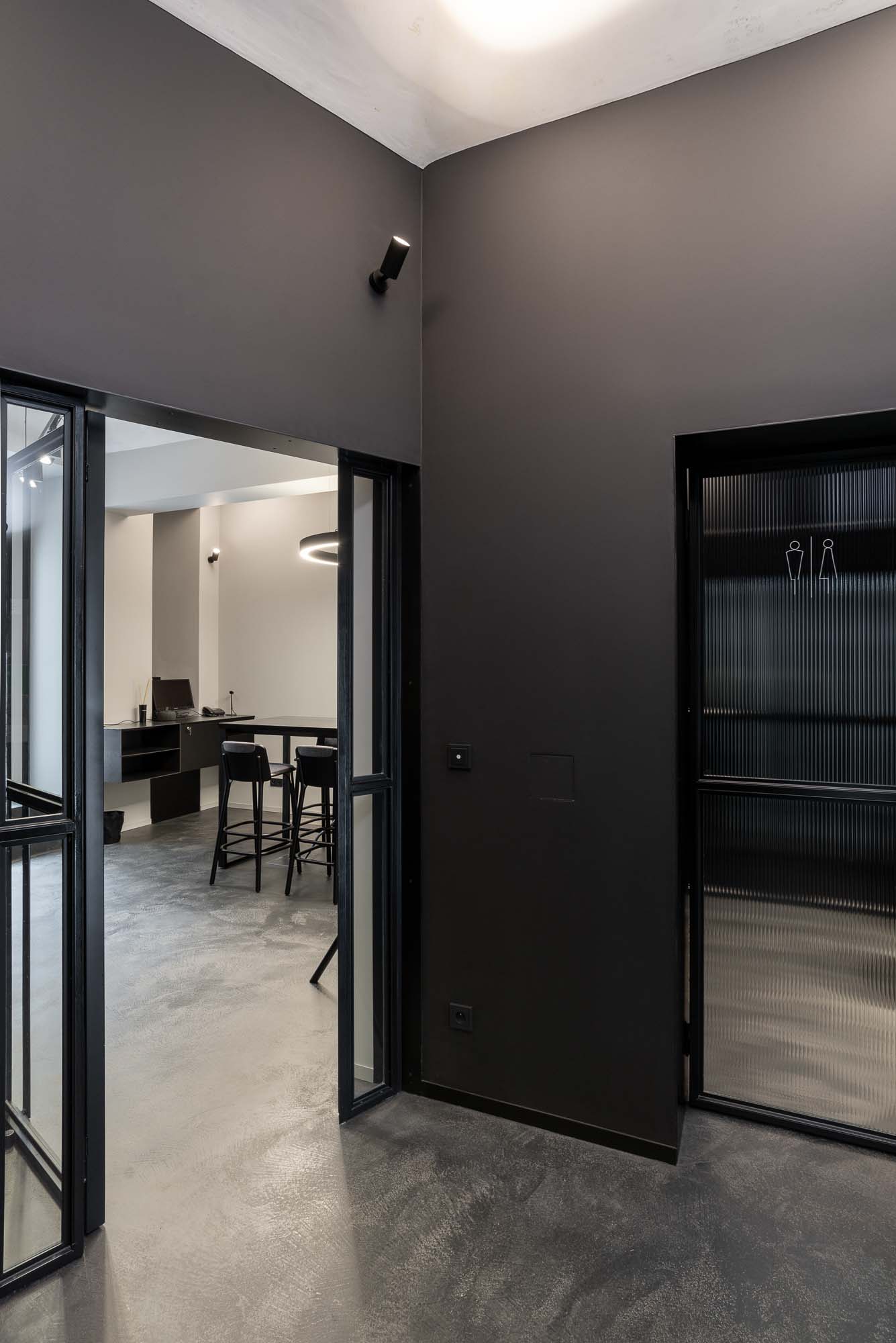Pohled do zasedací místnosti kanceláří Vanguard přes prosklenou stěnu v černém rámu, betonová stěrka na podlaze a minimalistický industriální interiér. Design by OOOOX