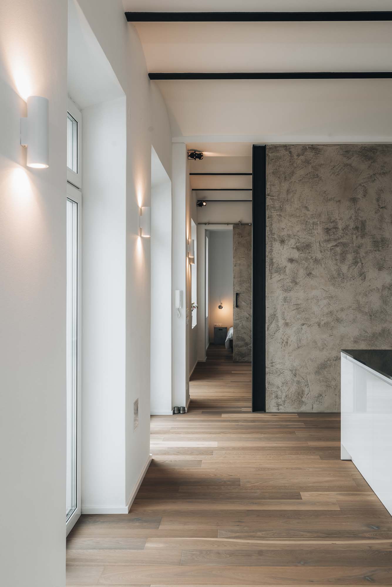 Detail otevřeného prostoru kuchyně s dřevěnou podlahou, dekorativní štukovou omítkou na stěně a liniovými designovými svítidly v minimalistickém interiéru. Design by OOOOX
