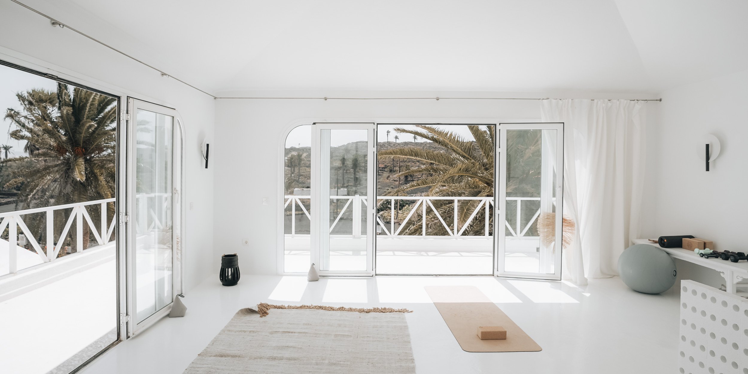Yoga místnost s bílými stěnami, otevřenými skládacími dveřmi na terasu s výhledem na palmy, korkový podložka, cvičební míč. OOOOX
