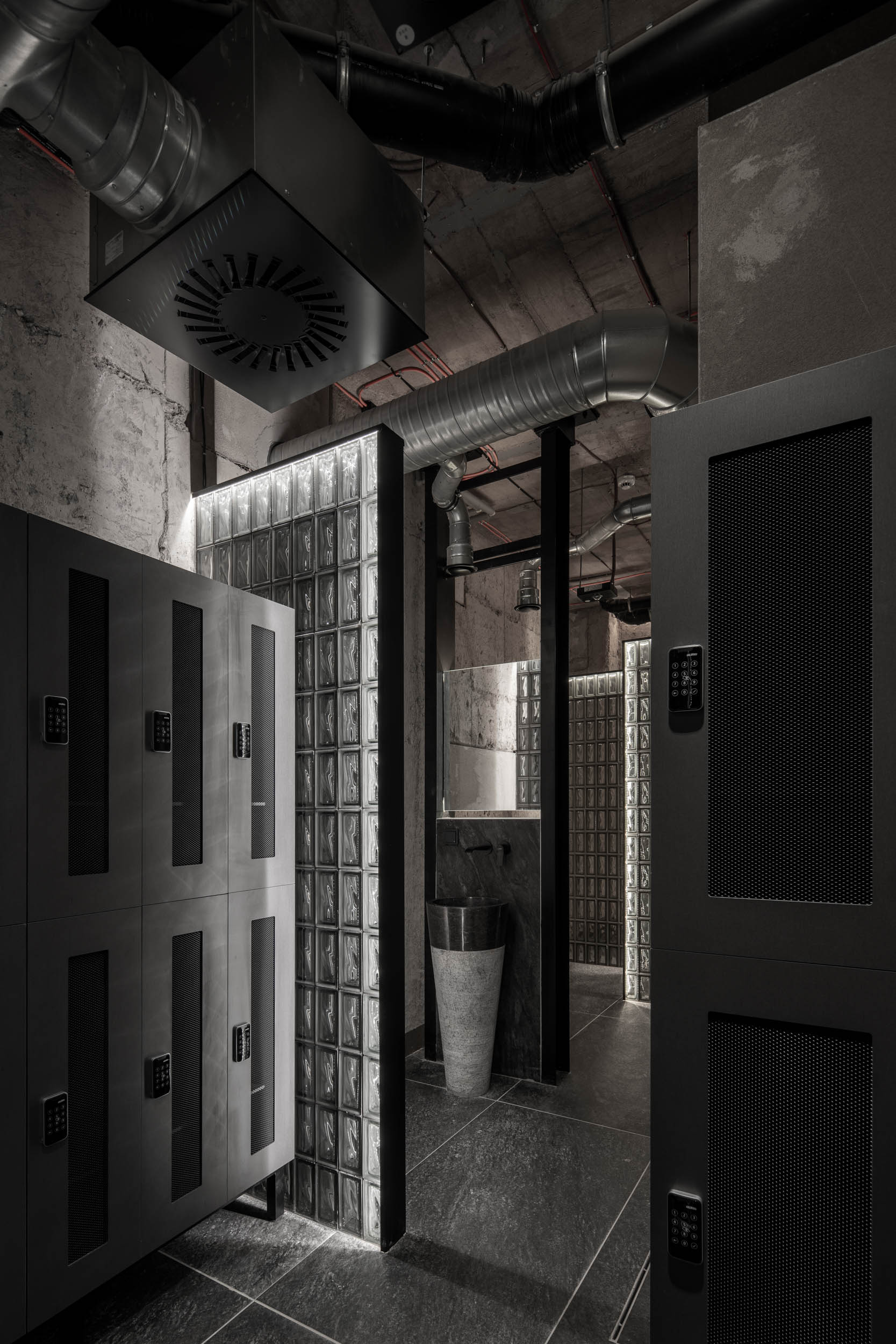 VANGUARD wellness locker room — dark lockers with digital locks, illuminated glass block partitions, stone pedestal sink, exposed ceiling pipes | OOOOX