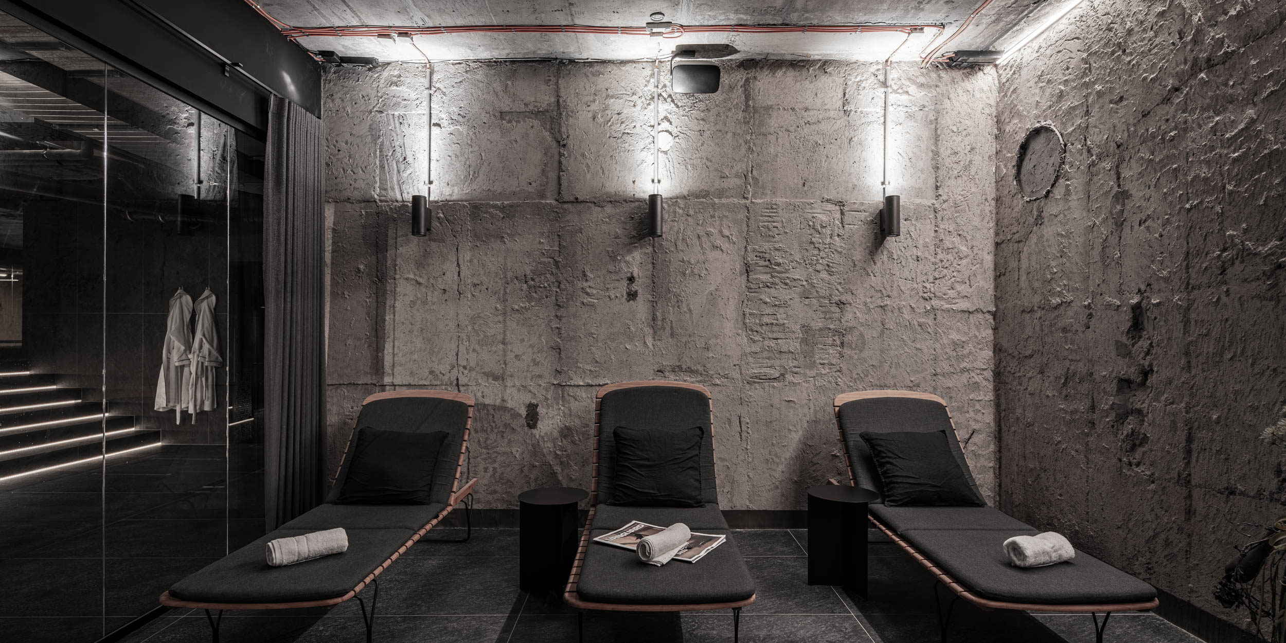 VANGUARD wellness relaxation lounge — three dark designer sun loungers by raw concrete bunker wall, ambient lighting, white robes | OOOOX