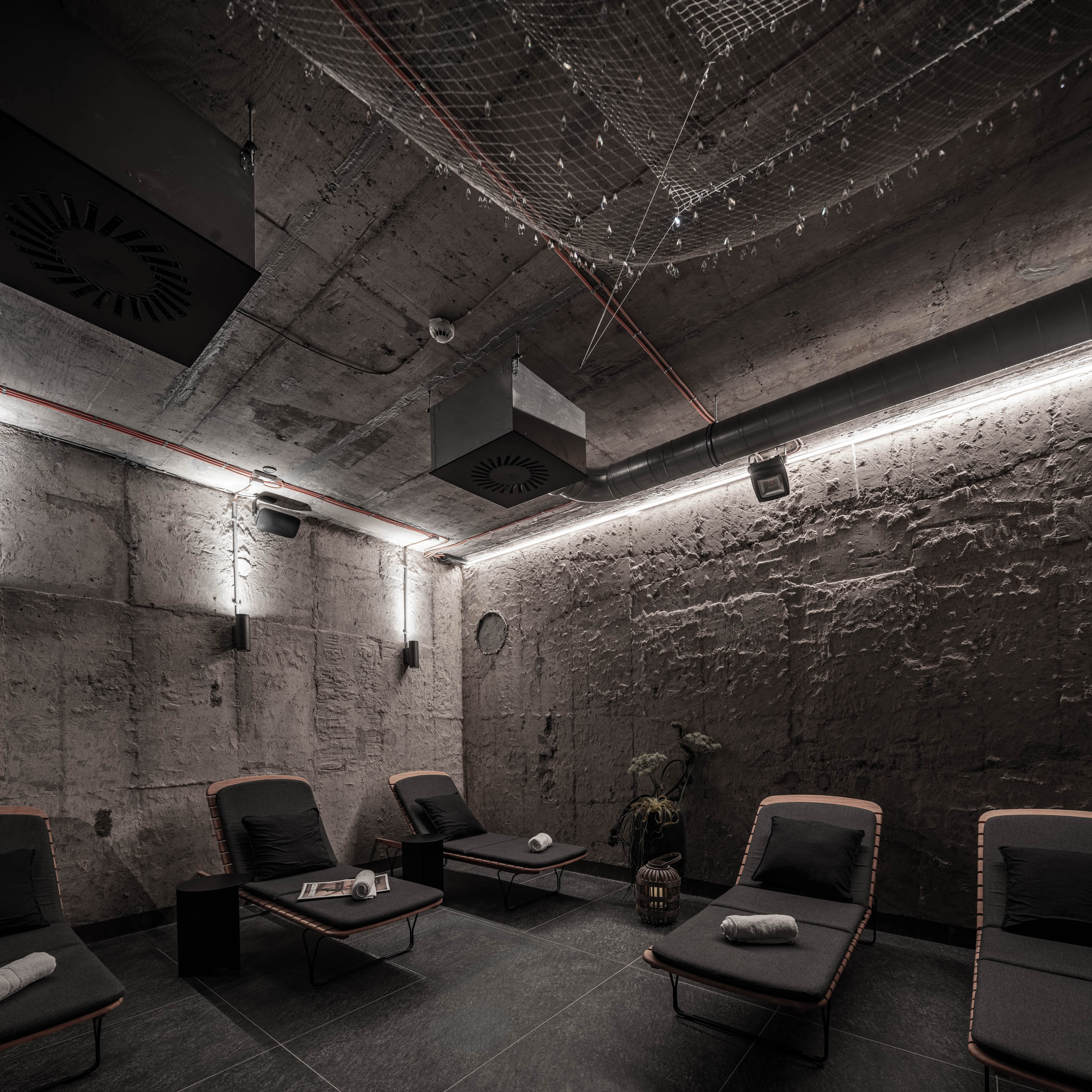 VANGUARD wellness relaxation lounge — overview with dark loungers in converted bunker, fishing net ceiling light with crystals, exposed pipes | OOOOX