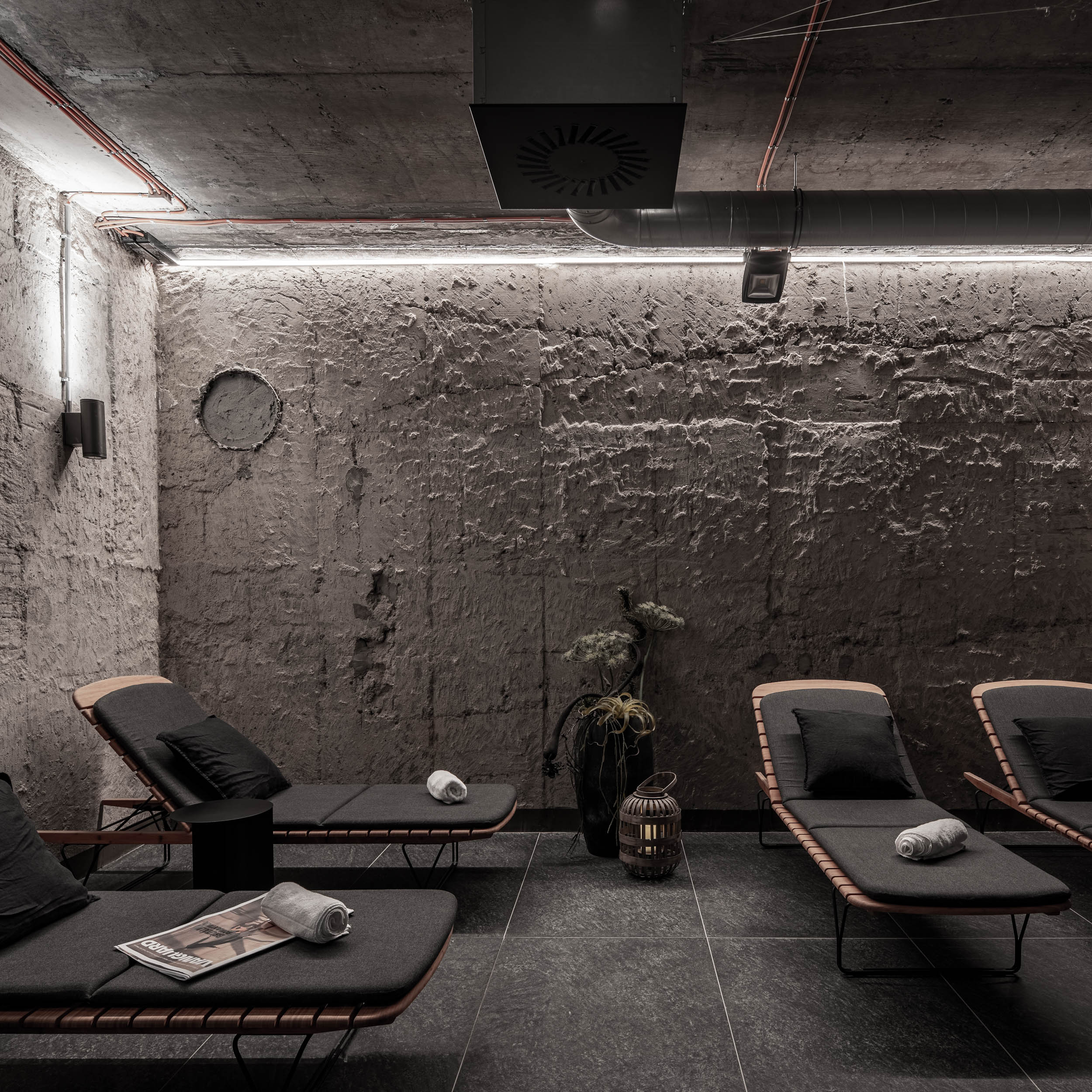 VANGUARD wellness relaxation lounge — dark loungers with cushions, raw textured concrete bunker walls, exposed pipes, decorative lantern | OOOOX