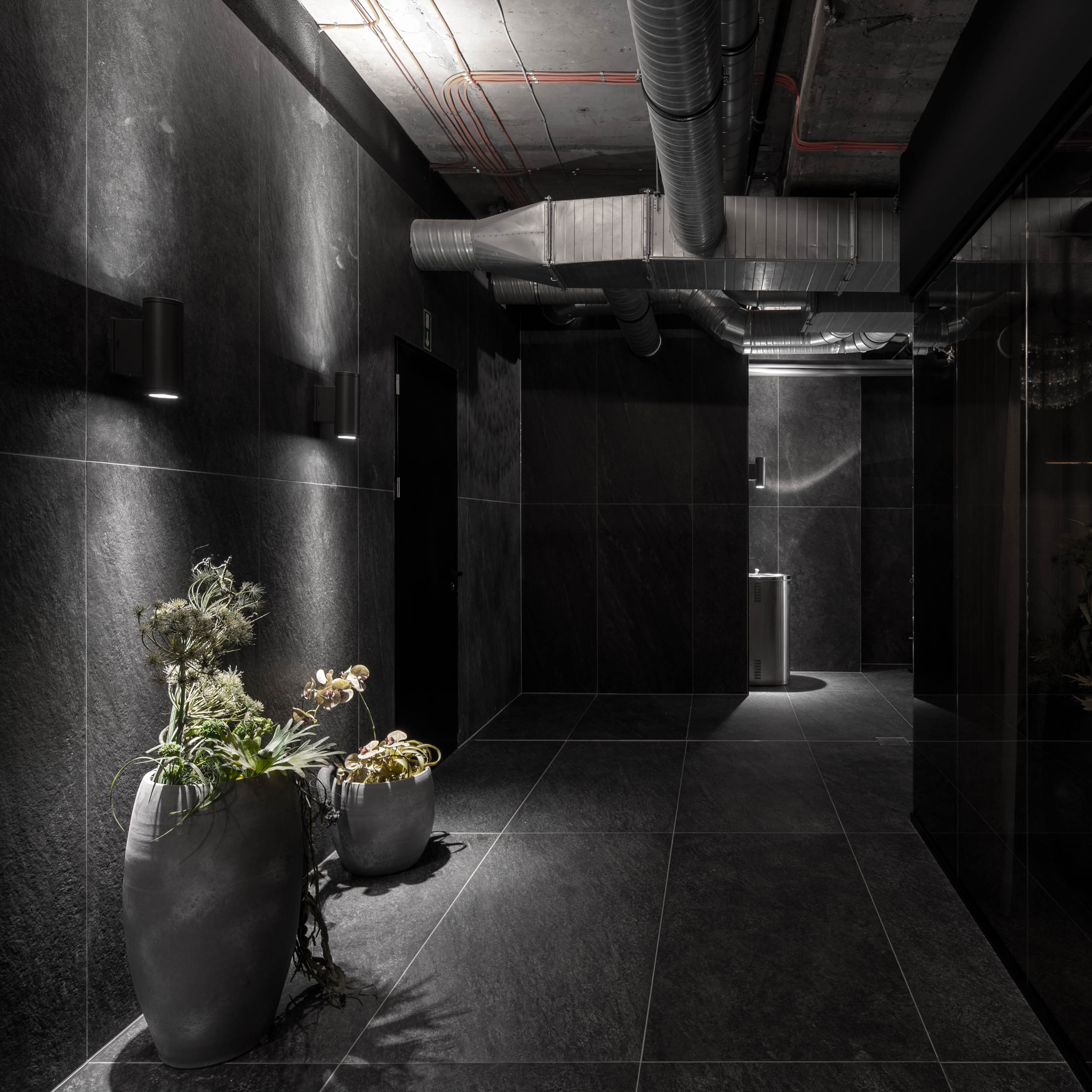 VANGUARD wellness corridor — dark hallway with black stone cladding, large planting vessels, exposed ductwork, industrial atmosphere | OOOOX