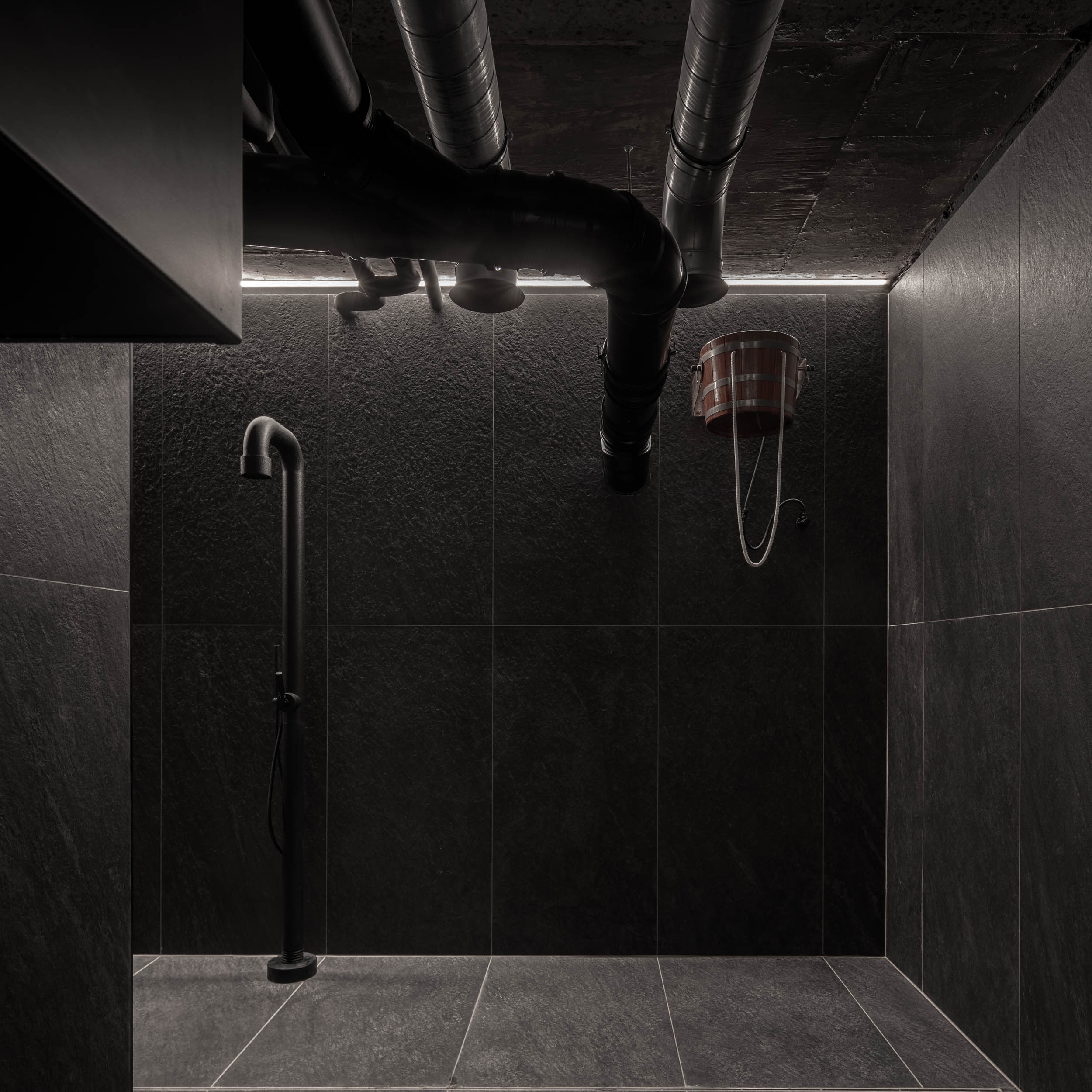 VANGUARD wellness shower — black shower column on dark tile, exposed ceiling pipes, copper ice water bucket, industrial design | OOOOX