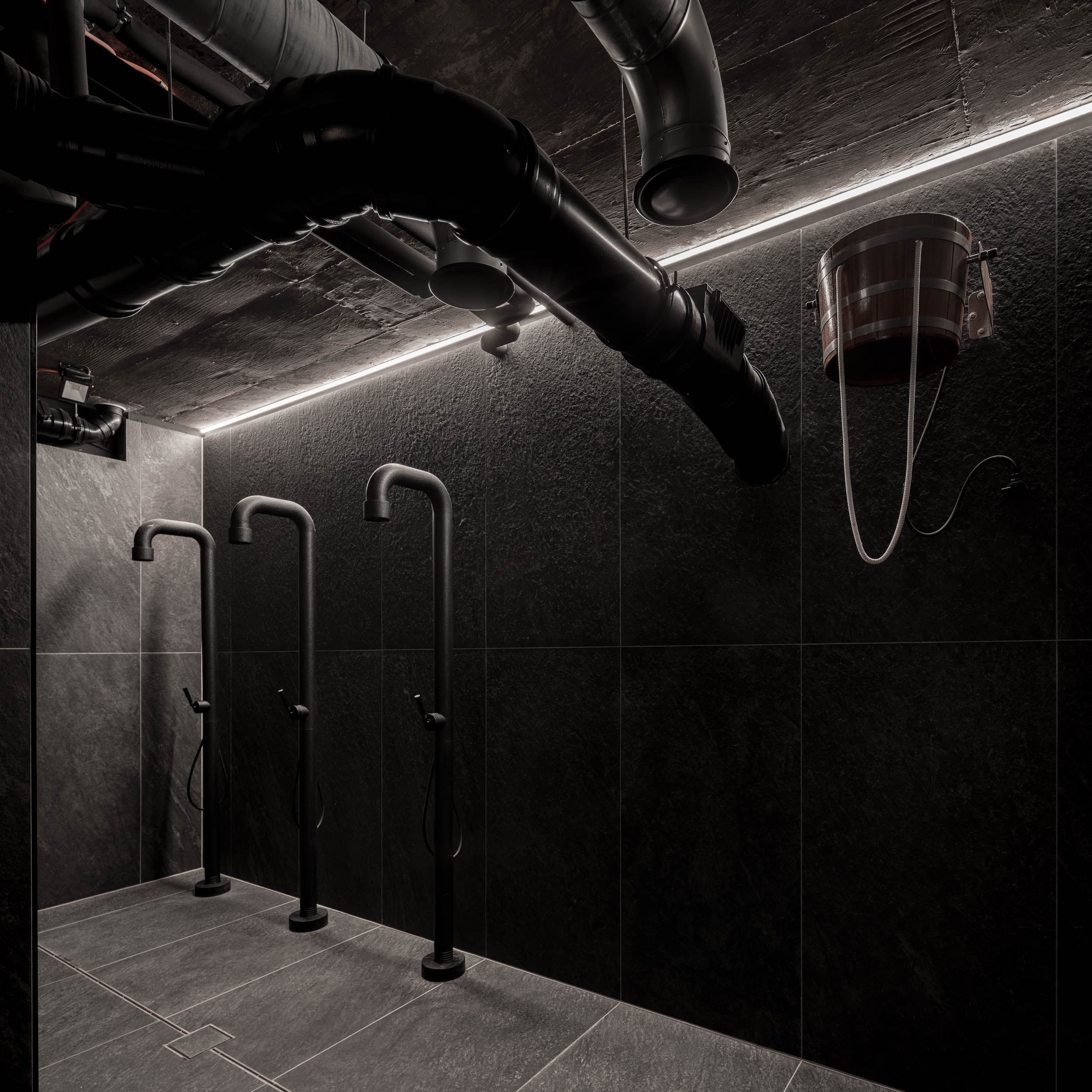 VANGUARD wellness showers — three black shower columns in dark-tiled shower room, exposed ceiling pipes, copper ice water bucket | OOOOX