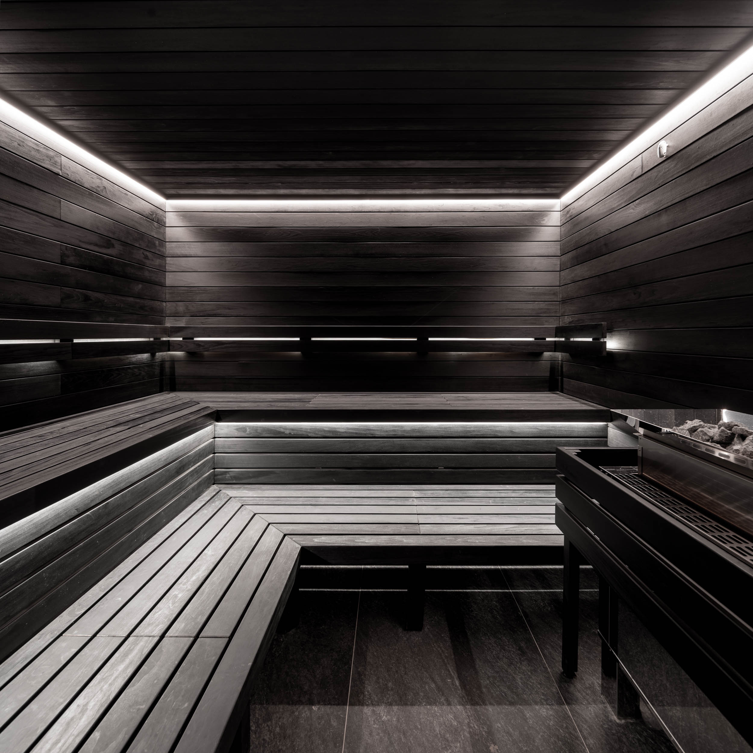 VANGUARD wellness Finnish sauna — view of sauna heater with stones, dark-stained wooden benches, LED strip light, minimalist industrial design | OOOOX