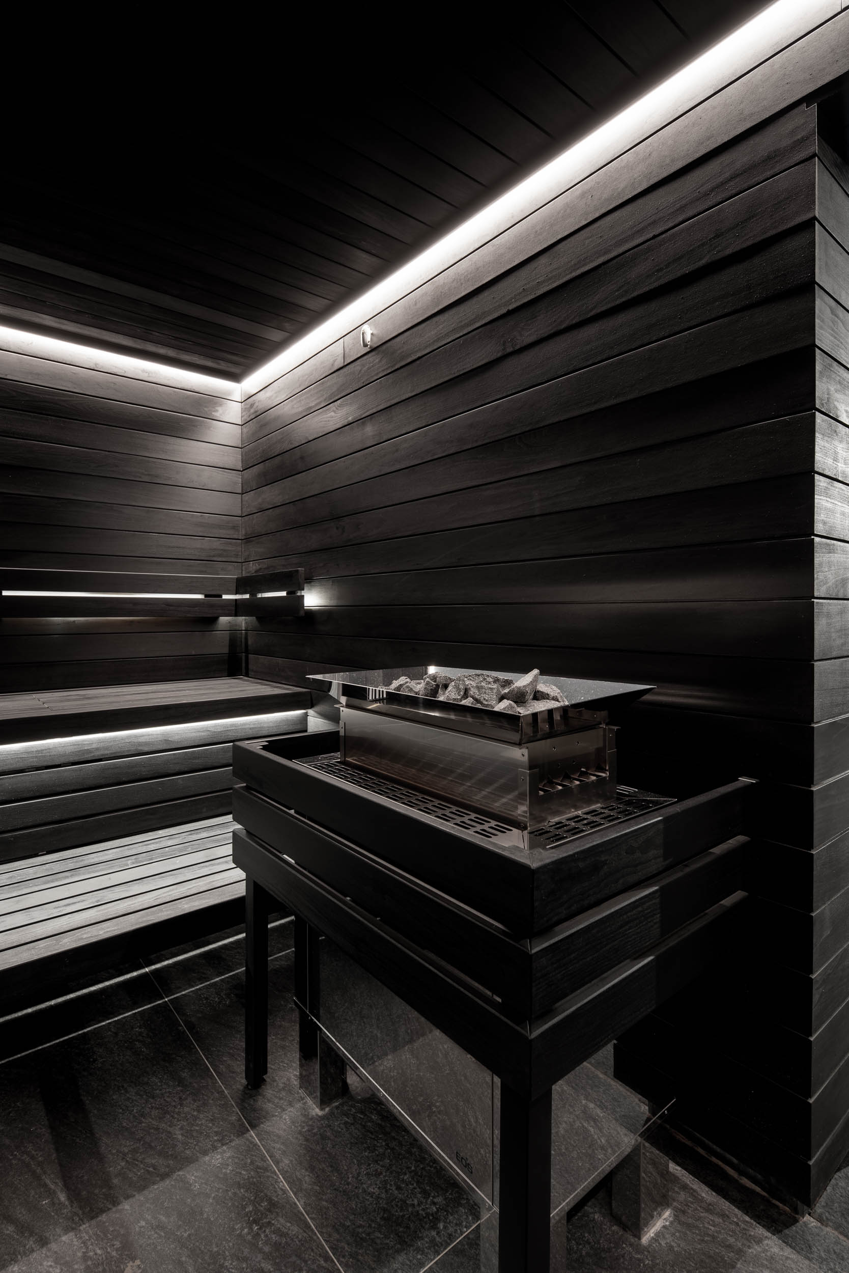 VANGUARD wellness Finnish sauna — detail of sauna heater with stones, dark-stained wood benches with LED underlight, black minimalist interior | OOOOX