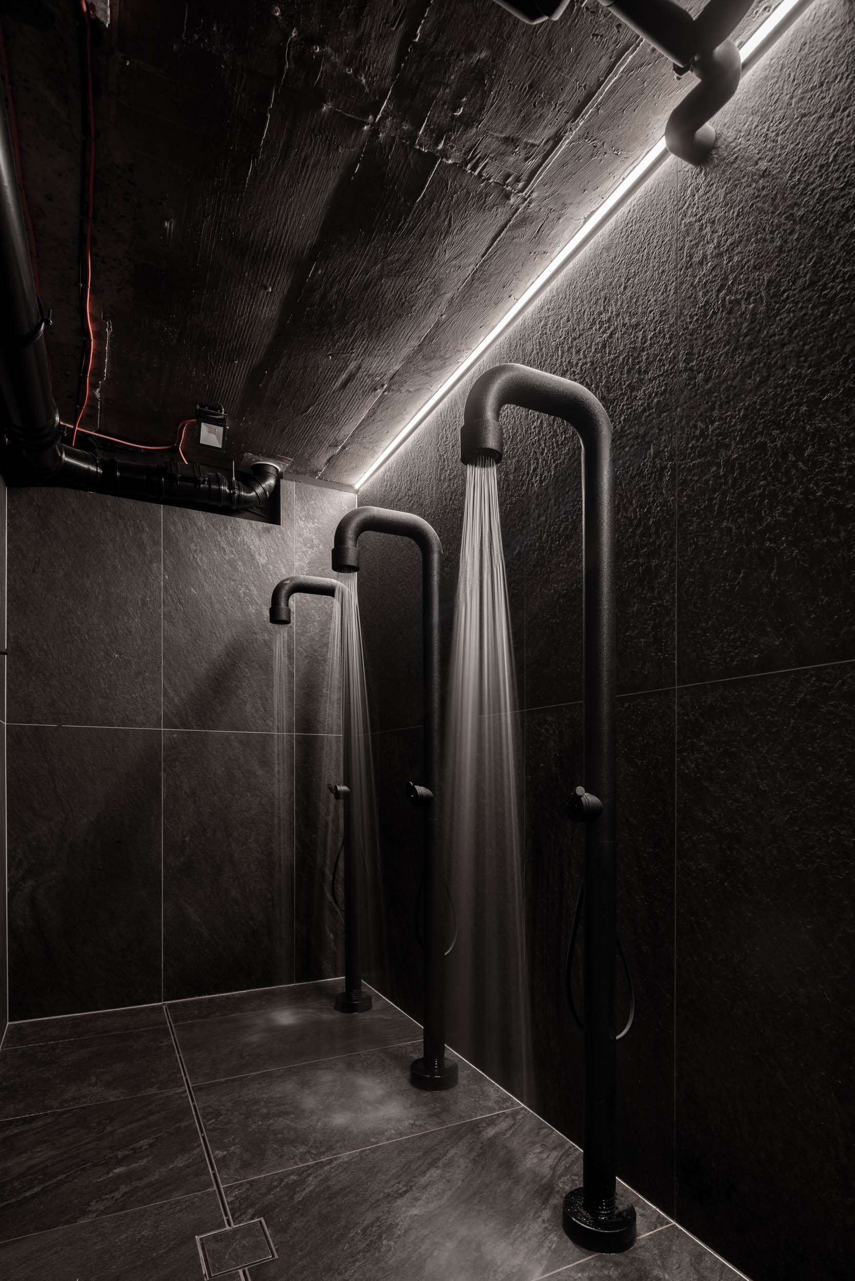 VANGUARD wellness active showers — water flowing from black shower column, exposed pipes on concrete ceiling, dark tile, industrial atmosphere | OOOOX