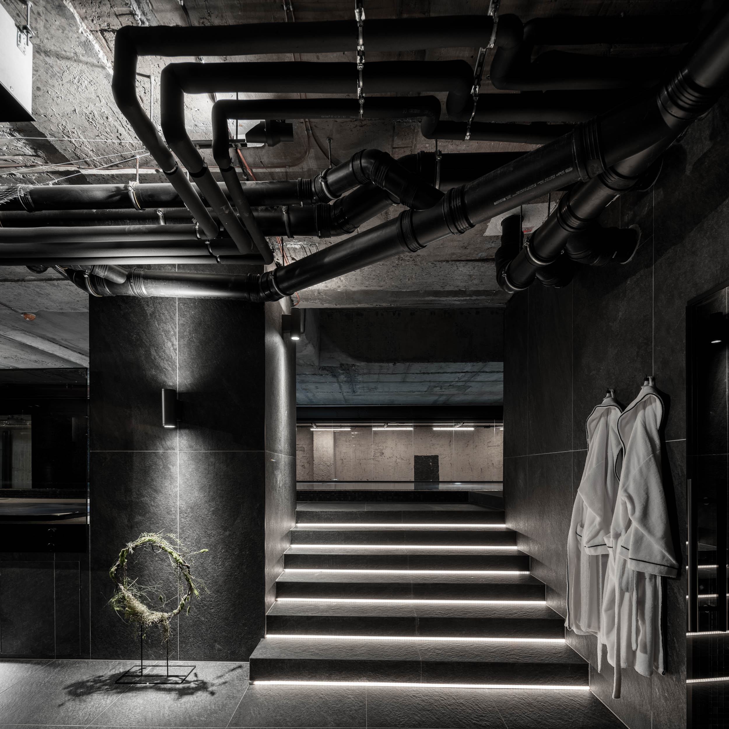 VANGUARD wellness entrance area — exposed pipes on bunker concrete ceiling, LED-lit staircase, white robes on hooks, decorative wreath installation | OOOOX