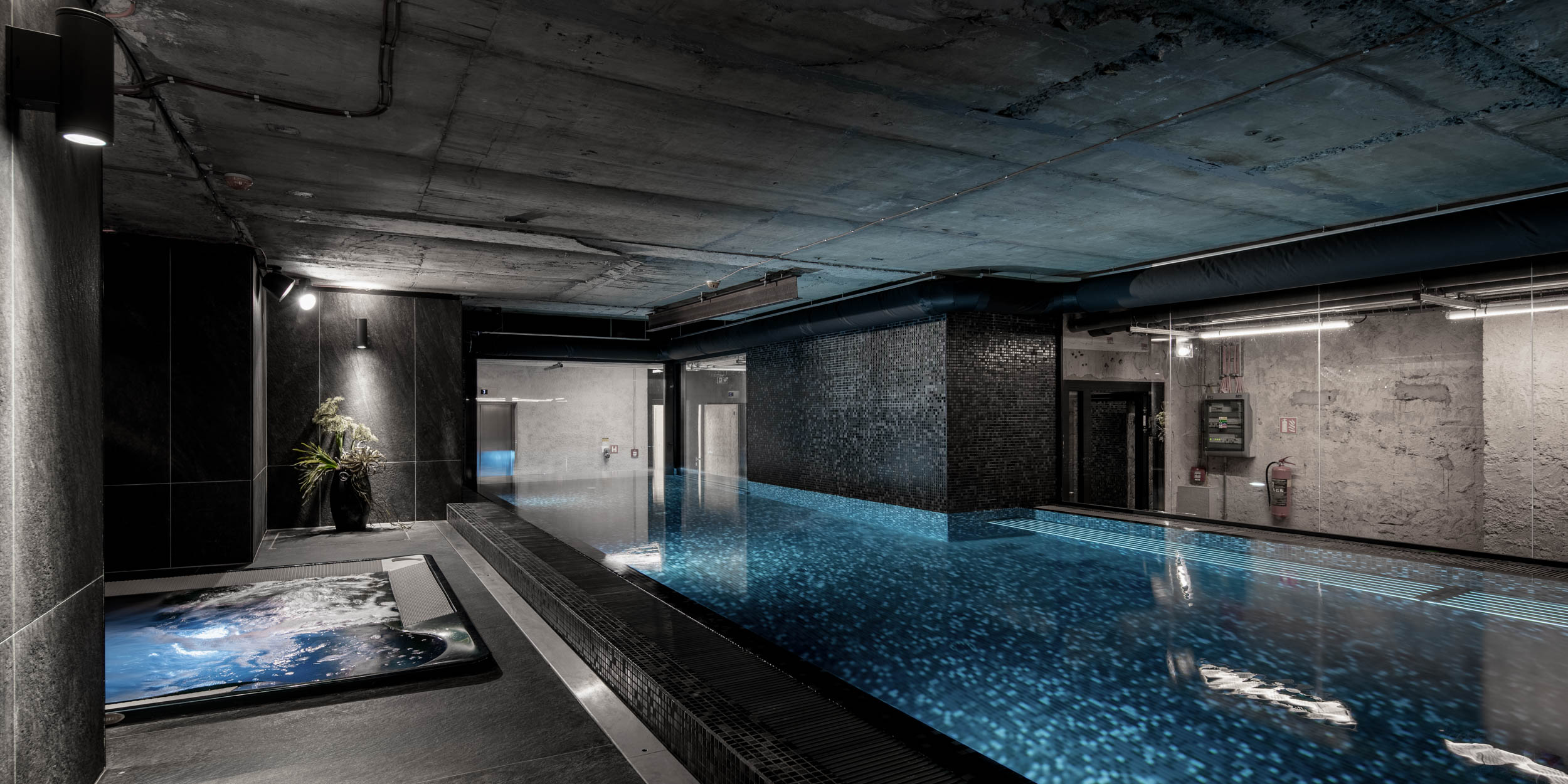 VANGUARD wellness pool — overview of lap pool with black mosaic and hot tub, raw concrete civil-defense shelter ceilings, linear lighting | OOOOX