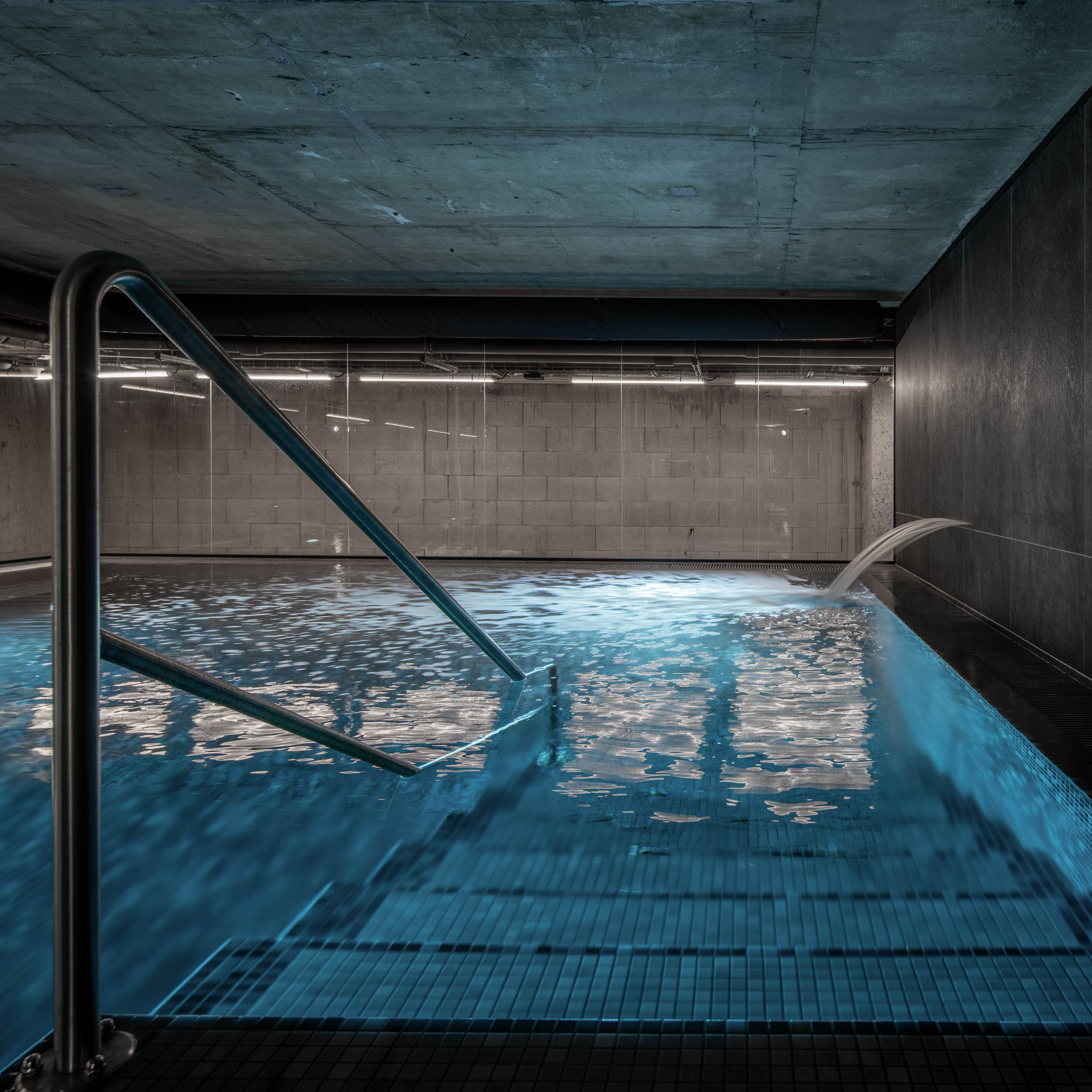 VANGUARD wellness pool — metal pool ladder into black mosaic pool, water jet from wall feature, linear lighting, concrete ceiling | OOOOX