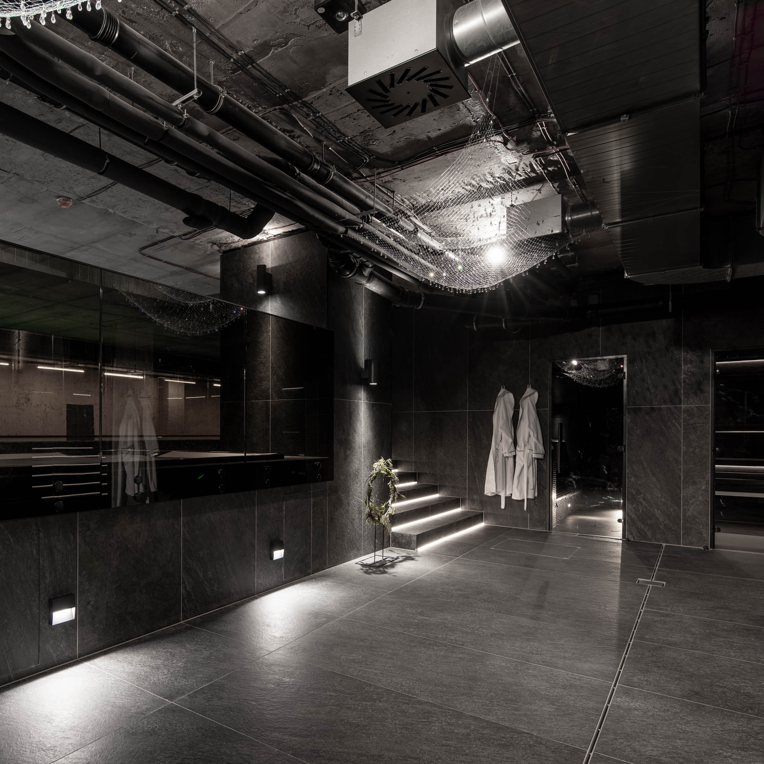 VANGUARD wellness entrance foyer — dark space with lit staircase, white robes, crystal-net chandelier, exposed pipes, industrial atmosphere | OOOOX