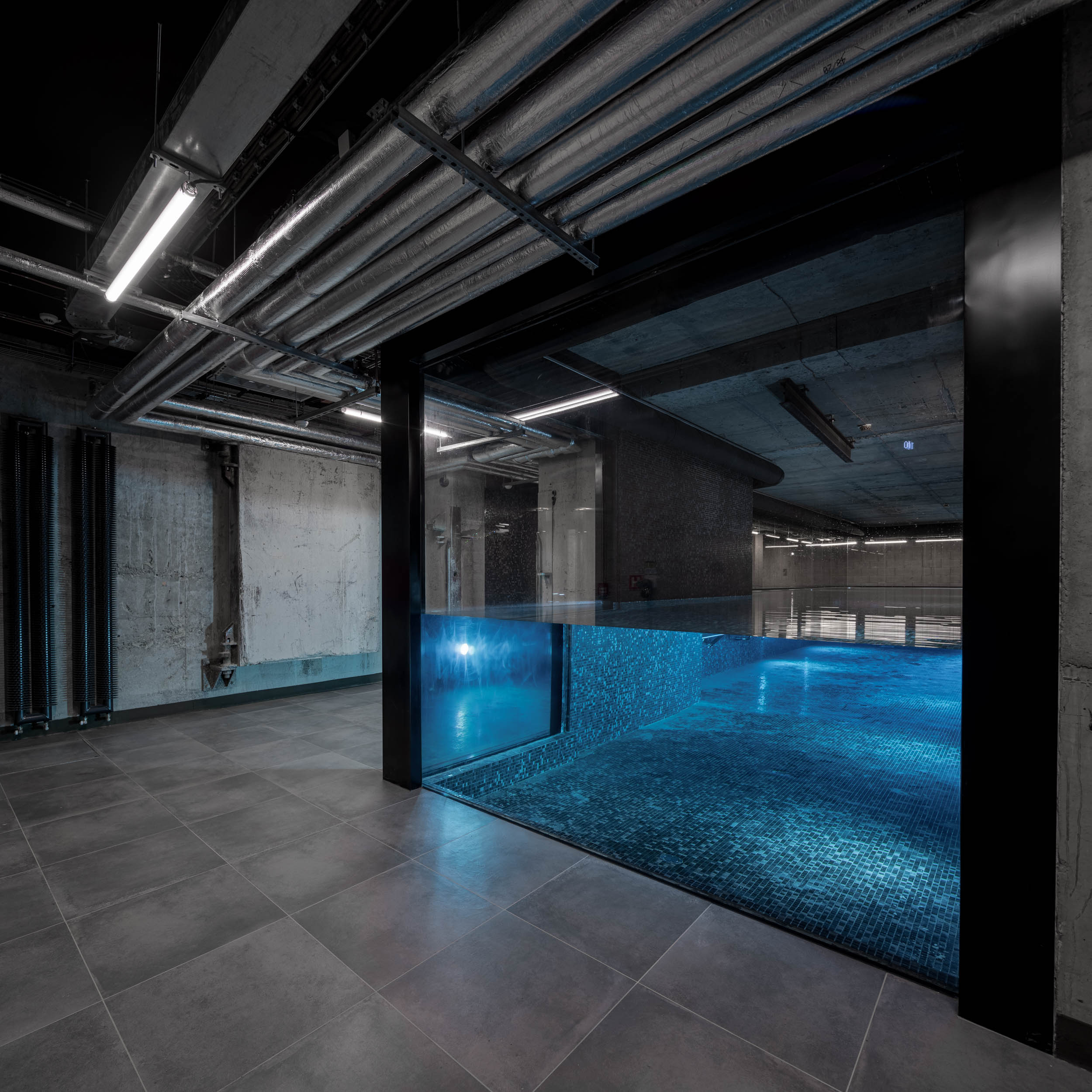 VANGUARD wellness elevator lobby with pool glass front — view from lobby to illuminated black mosaic pool behind glass wall, exposed ceilings | OOOOX
