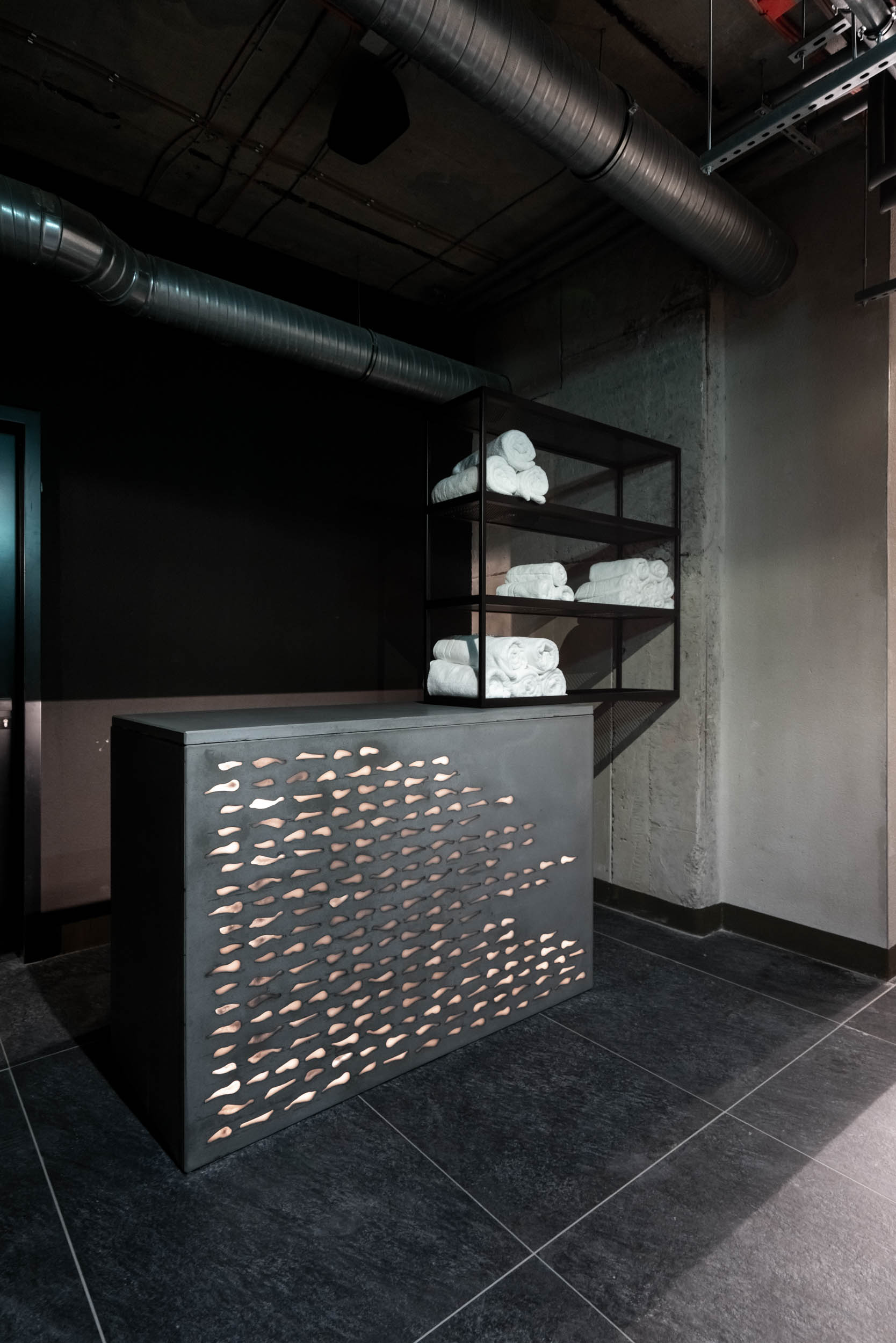 VANGUARD wellness reception desk — dark counter with laser-cut perforated steel front with organic pattern, shelving with white towels, exposed pipes | OOOOX