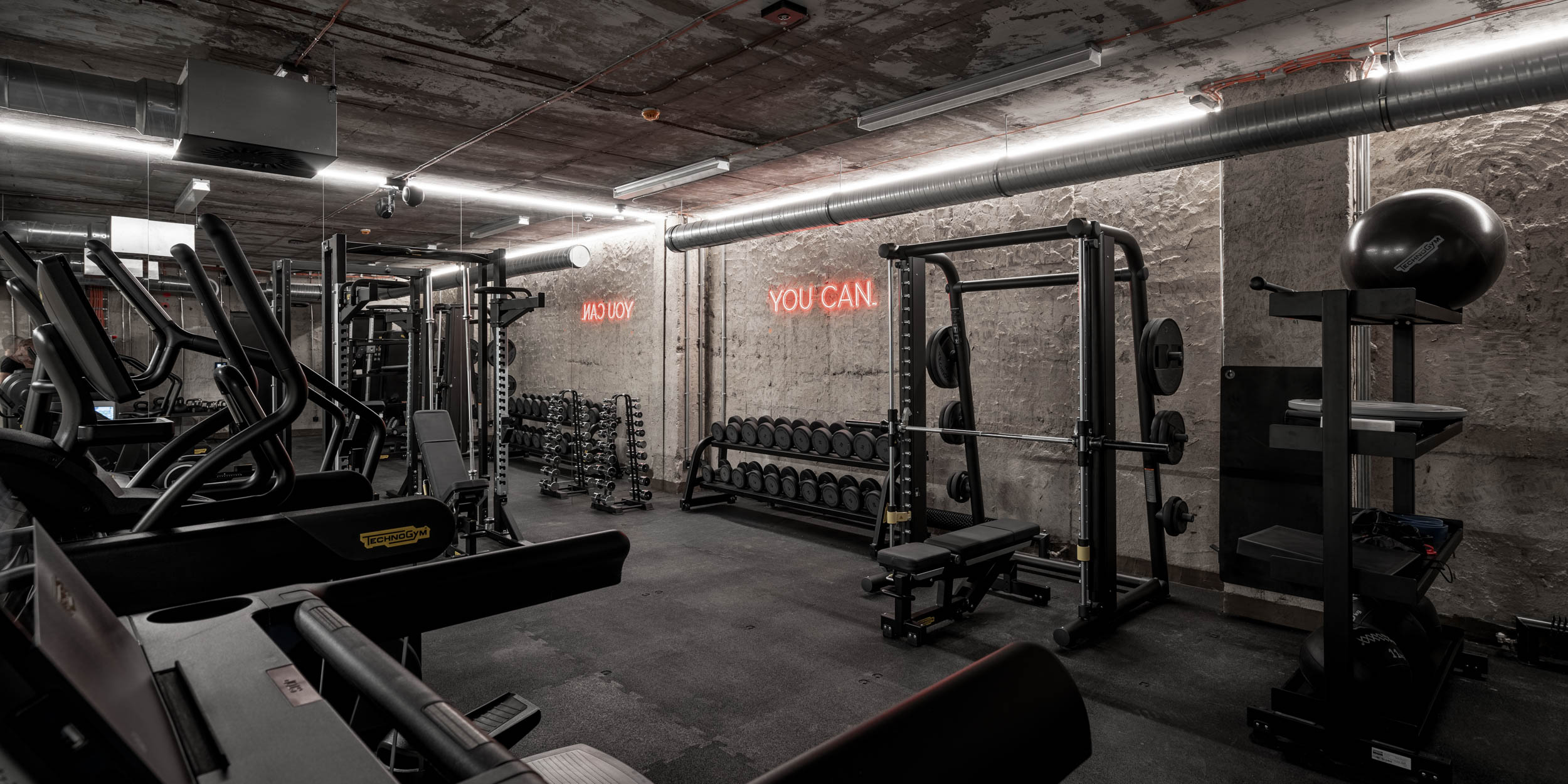 VANGUARD wellness gym — cardio and weight machines, dumbbell rack, neon motivational sign YOU CAN on raw concrete wall, industrial design | OOOOX