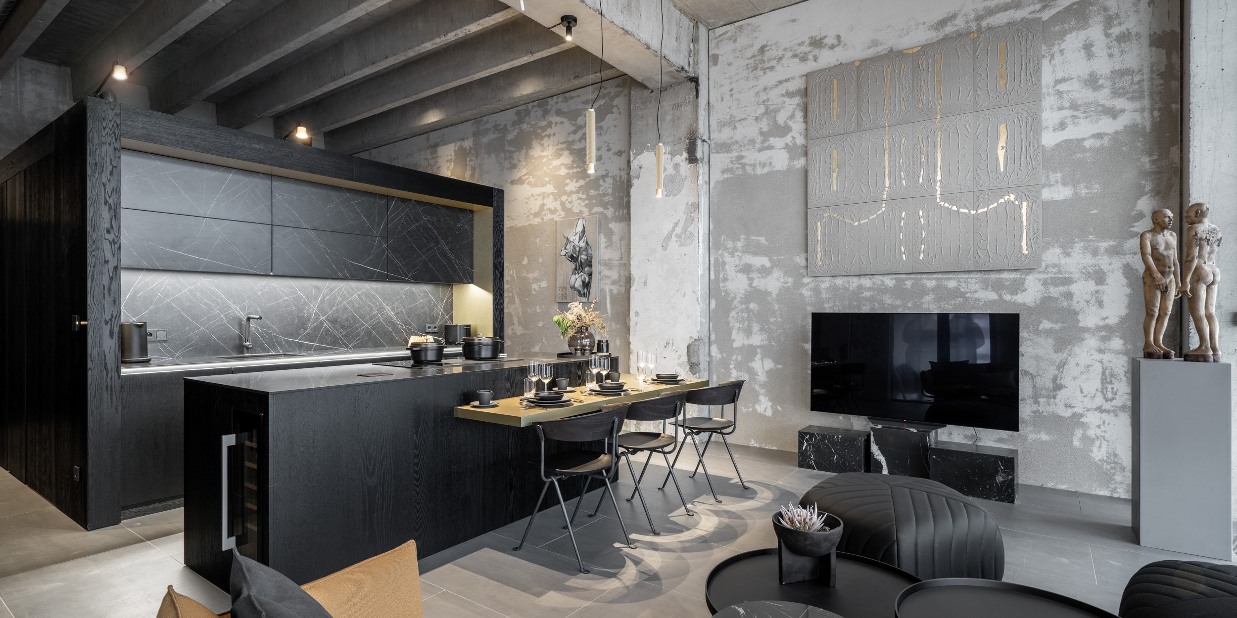 Full view of the Vanguard model loft – black kitchen cube with marble island and cantilevered dining table in brass metal laminate, living area in the background with textured artwork on raw concrete wall. OOOOX