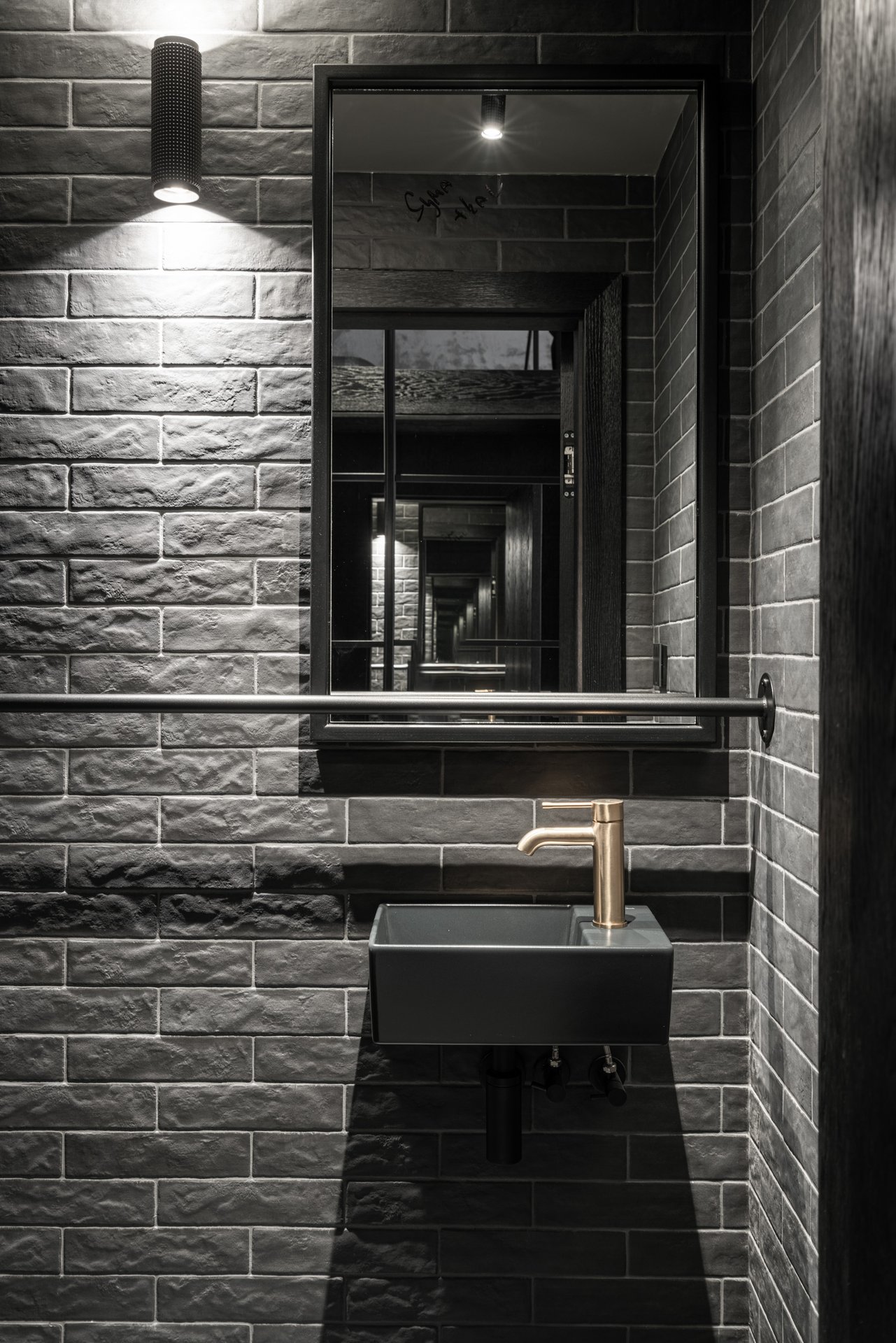 Kompaktní separátní WC v loftu Vanguard – malé tmavé kameninové umyvadlo s mosaznou baterií, černé cihlové obklady, černé zrcadlo, bodové osvětlení. OOOOX