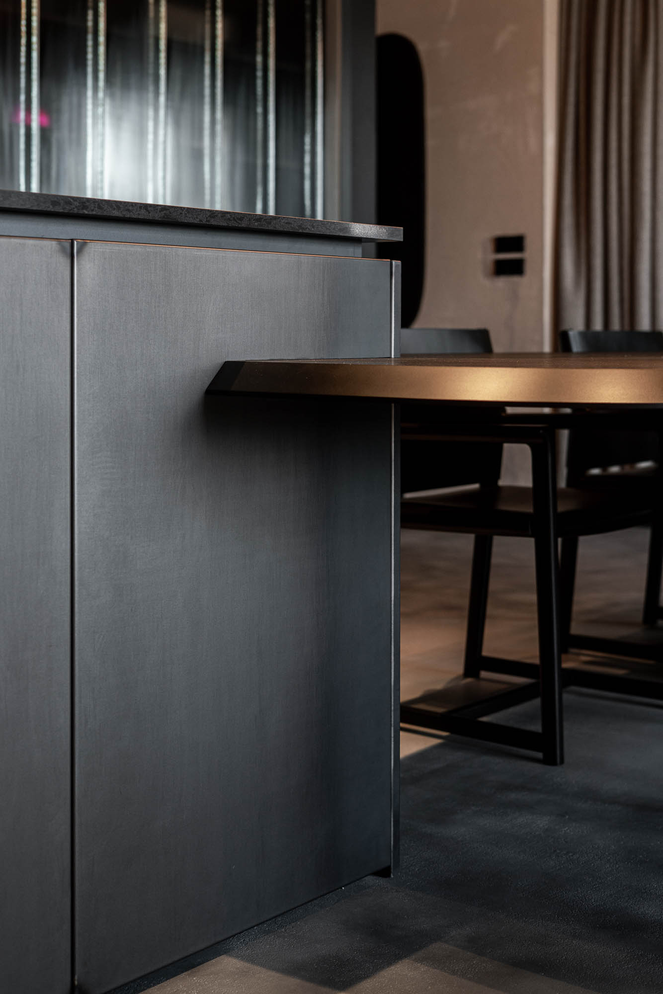 Detail napojení oválného černého jídelního stolu na kuchyňský ostrůvek v minimalistickém industriálním interiéru s designovým závěsným svítidlem. Design by OOOOX