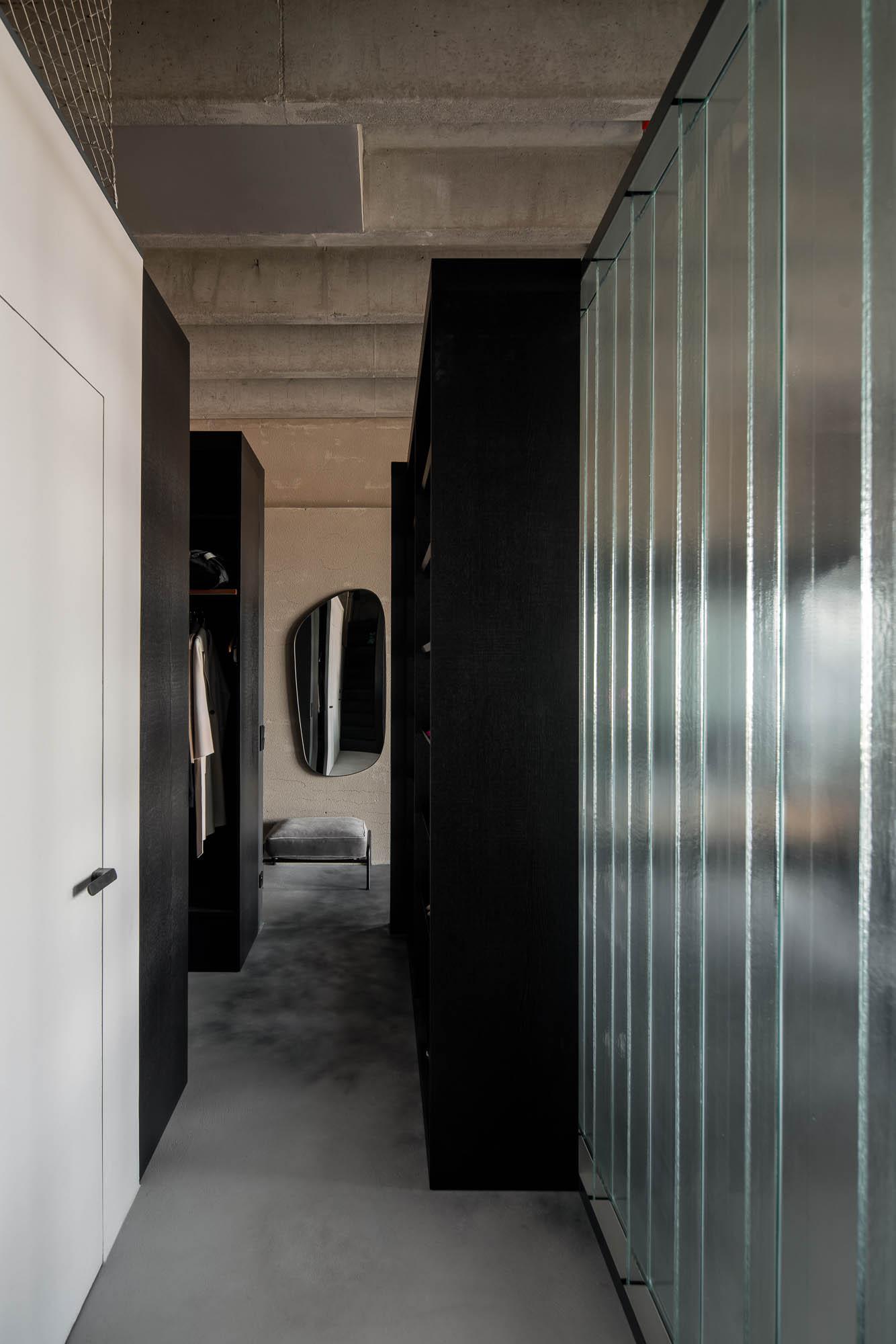 Pohled na soukromou chodbu oddělenou copilitem, betonovou stěrku na podlaze, odhalené betonové trámy, vestavěné šatní skříně a organicky tvarované designové zrcadlo. Design by OOOOX