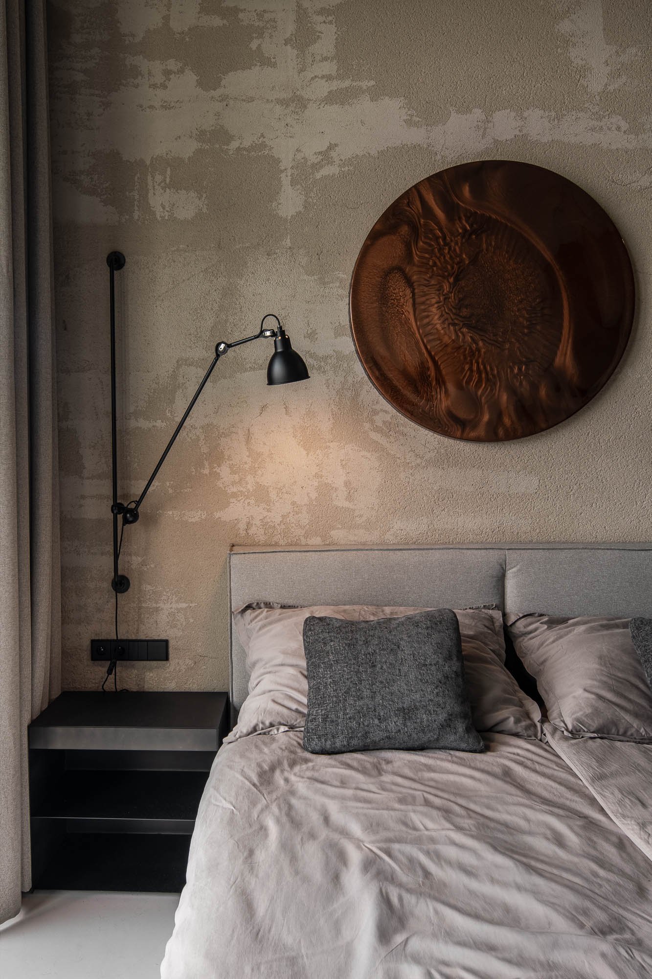 Noční stolek ložnice loftu Vanguard s rozsvícenou černou lampou, detail měděného kruhového díla a šedé lněné povlečení – OOOOX design