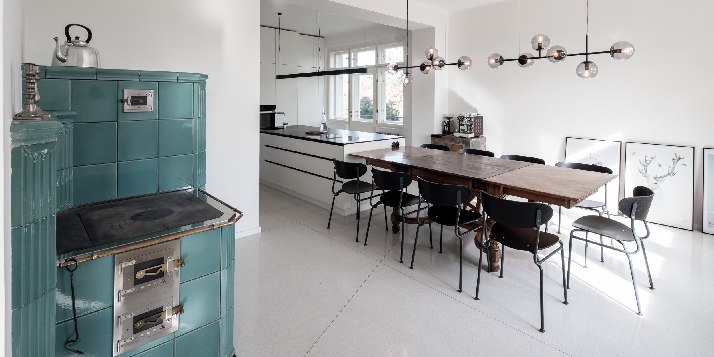 Dining room with kitchen – turquoise tiled stove on left, original carved table with black chairs, bubble glass pendant, white kitchen island, terrazzo floor. OOOOX