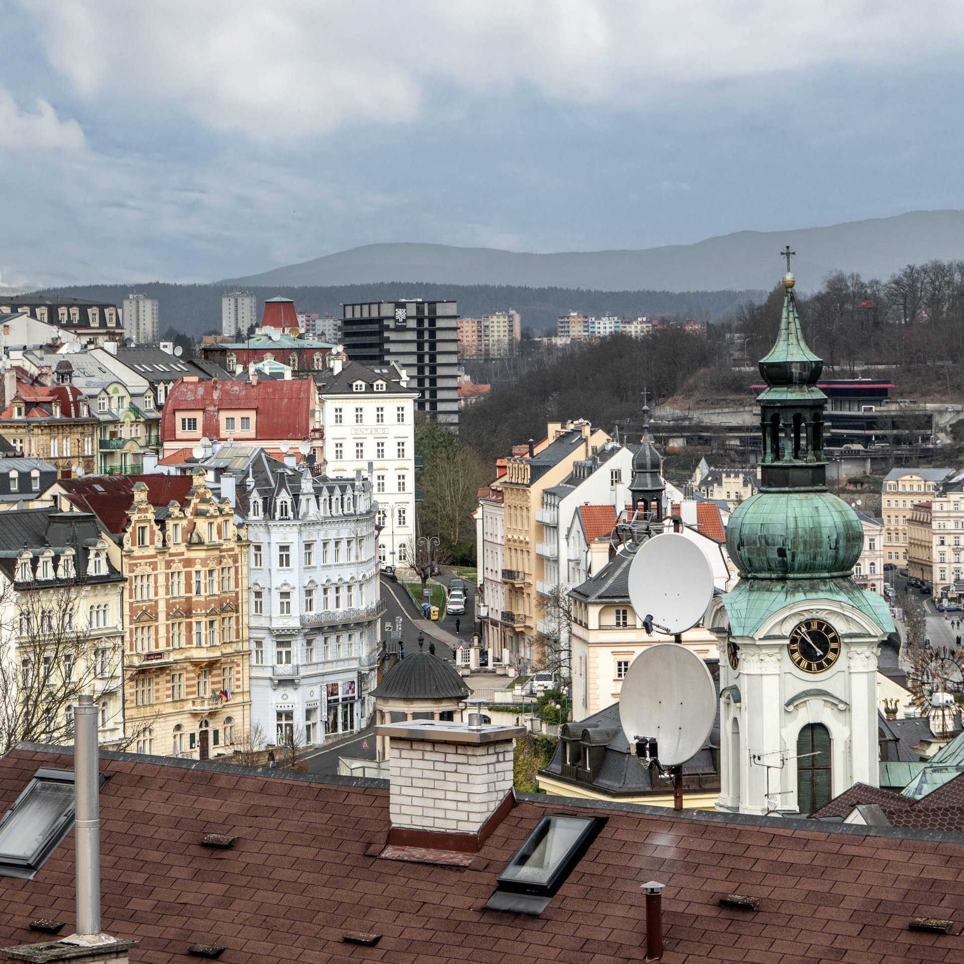 Výhled přes střechy Karlových Varů s věží kostela a okolními kopci | OOOOX