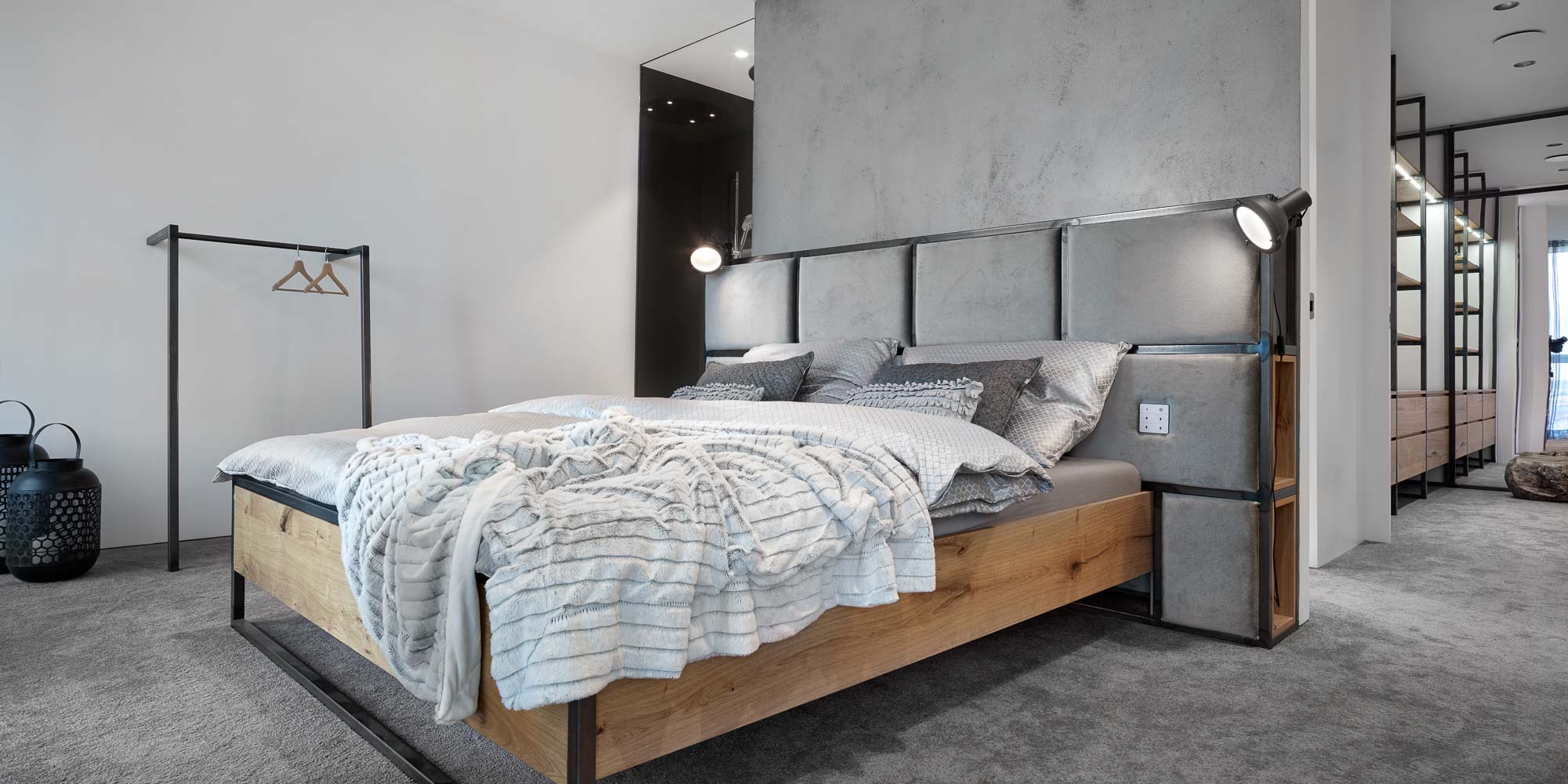 Bedroom of the V-Tower Prague apartment – bed with oak and steel frame, upholstered headboard with reading lamps, concrete plaster wall behind, open clothes rail and wardrobe shelving