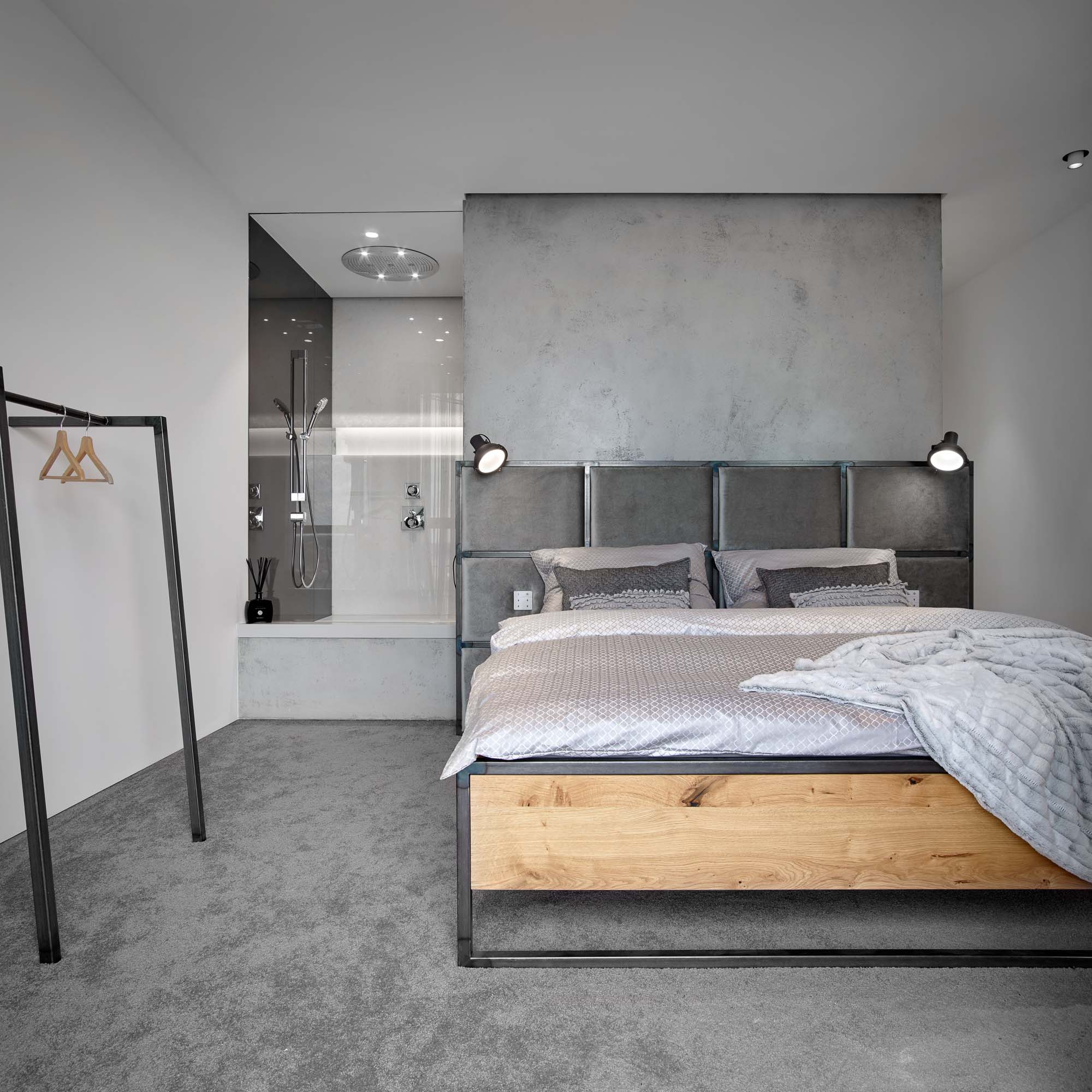 Bedroom of the V-Tower Prague apartment – full view of the oak-framed bed, upholstered headboard, walk-in shower with glass partition and rain shower head in the background, grey carpet