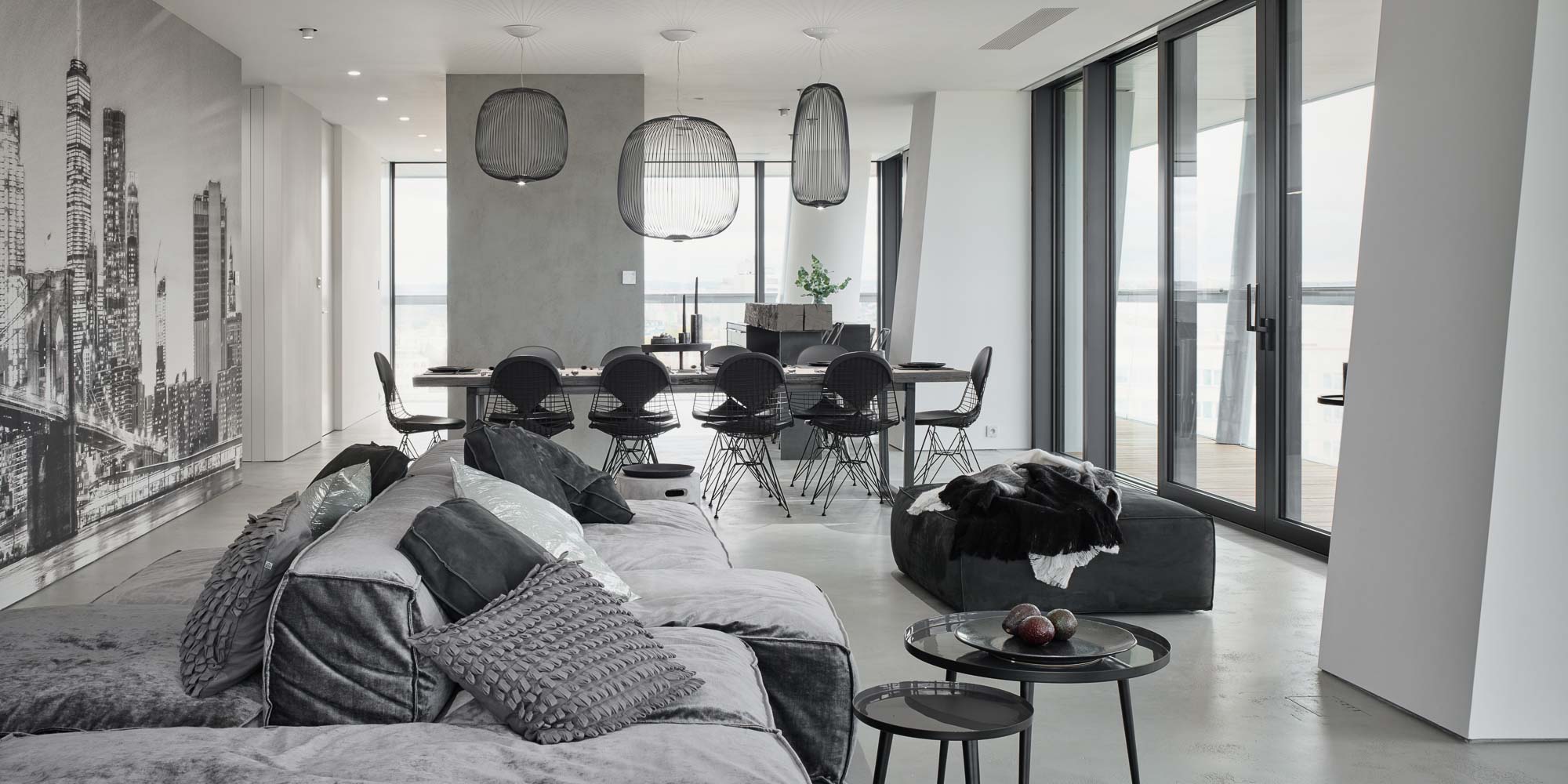 Open-plan living space in the V-Tower Prague apartment – grey sofa, dining table with wire chairs, rattan pendant lights, NYC skyline mural on the wall and panoramic windows overlooking Prague