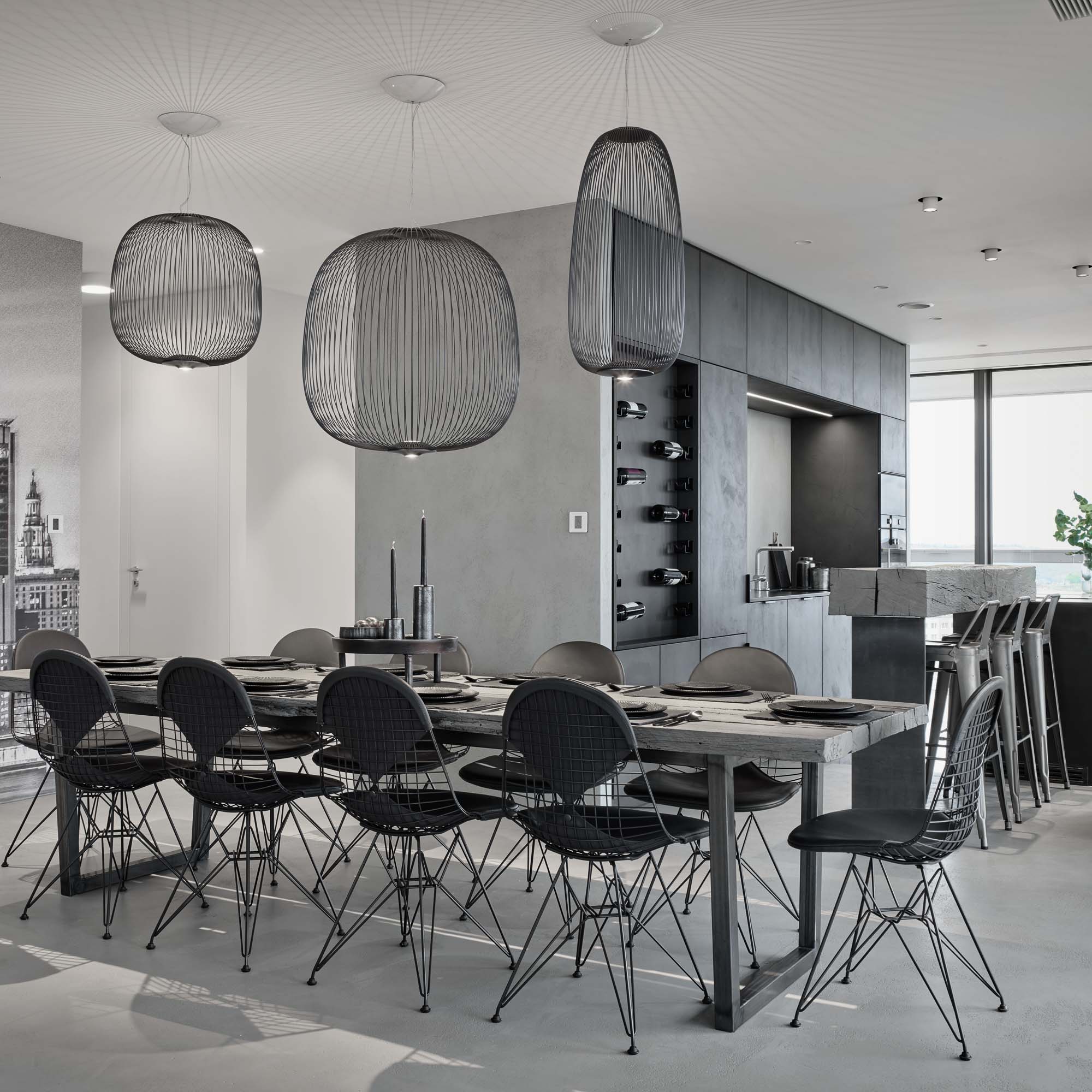 Dining area of the V-Tower Prague apartment – dark dining table with wire chairs, rattan pendant lights (Foscarini Spokes), dark kitchen cabinetry with wine rack niche, stone bar counter with metal bar stools