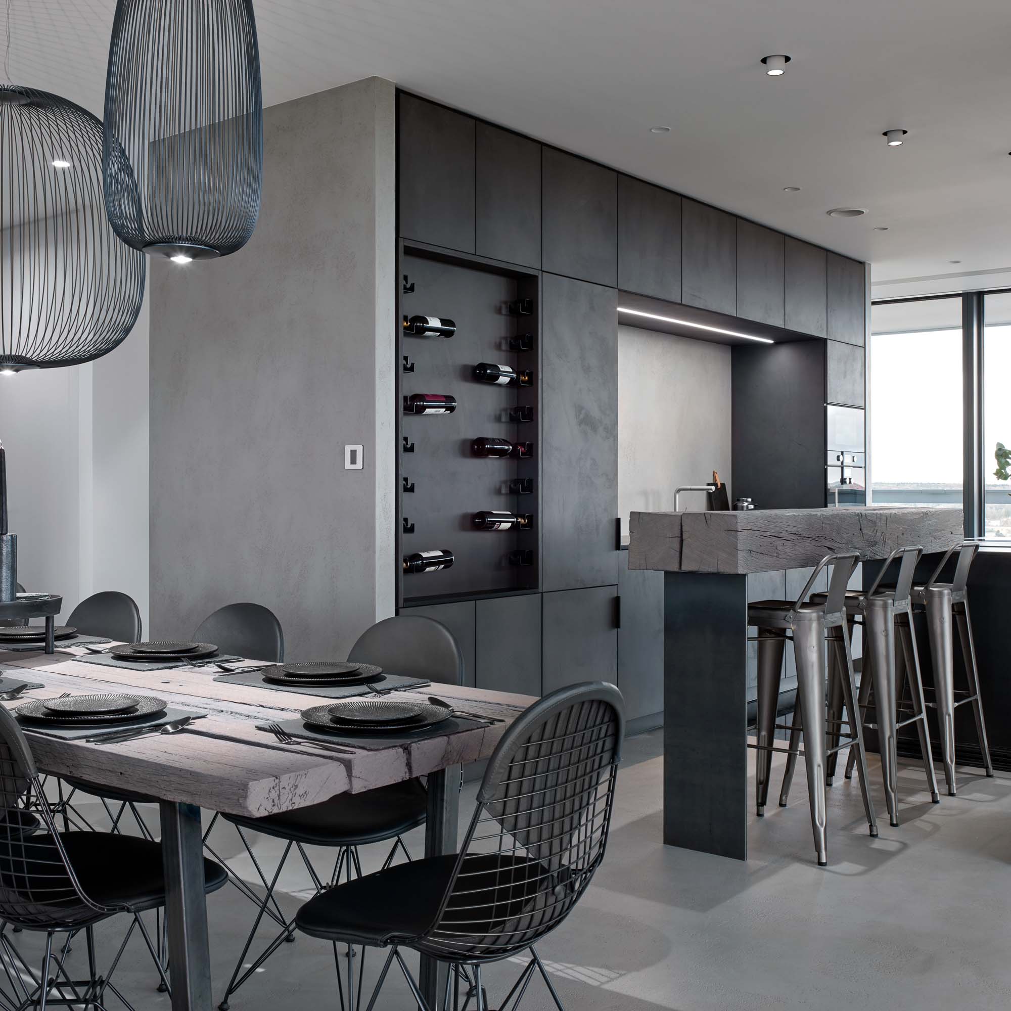 Jídelna a kuchyně bytu V-Tower Praha – detail jídelního stolu a drátěných židlí, tmavá zástavba s integrovaným vinným regálem, kamenný barový pult s kovovými stoličkami Tolix