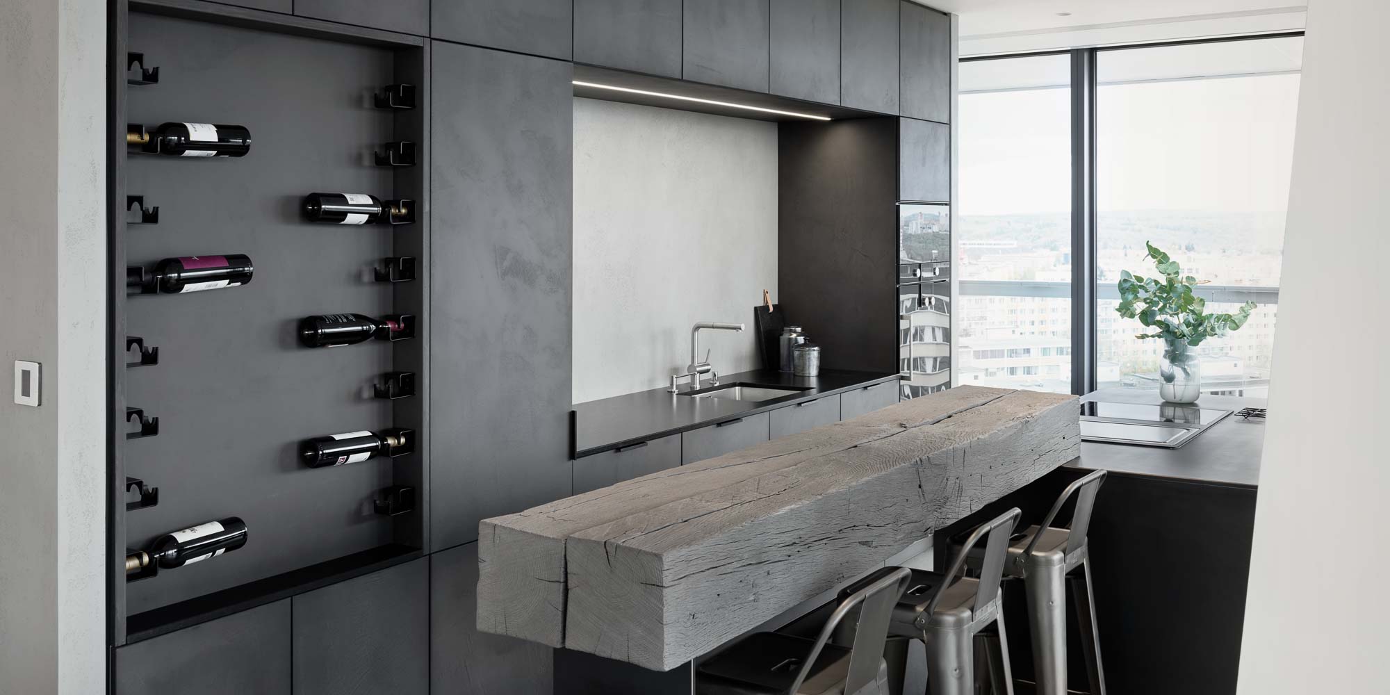 Kitchen of the V-Tower Prague apartment with city view – dark cabinetry with plaster finish, integrated wine rack, solid reclaimed wood bar counter, Tolix metal bar stools and panoramic Prague view through the window