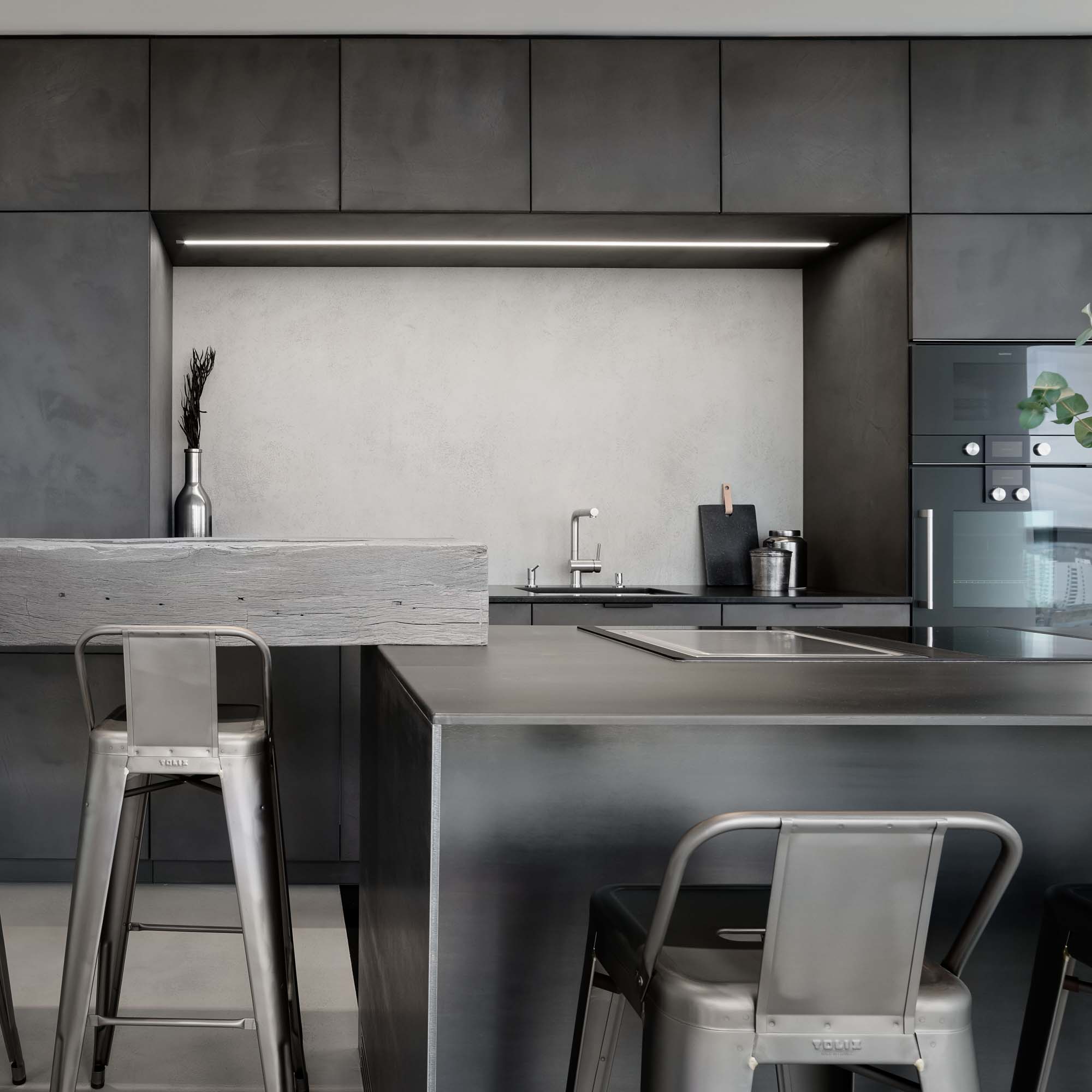 Kitchen detail of the V-Tower Prague apartment – dark kitchen island with stone/plaster countertop, Tolix metal bar stools, reclaimed wood bar counter, built-in Gaggenau appliances and LED strip under the cabinetry