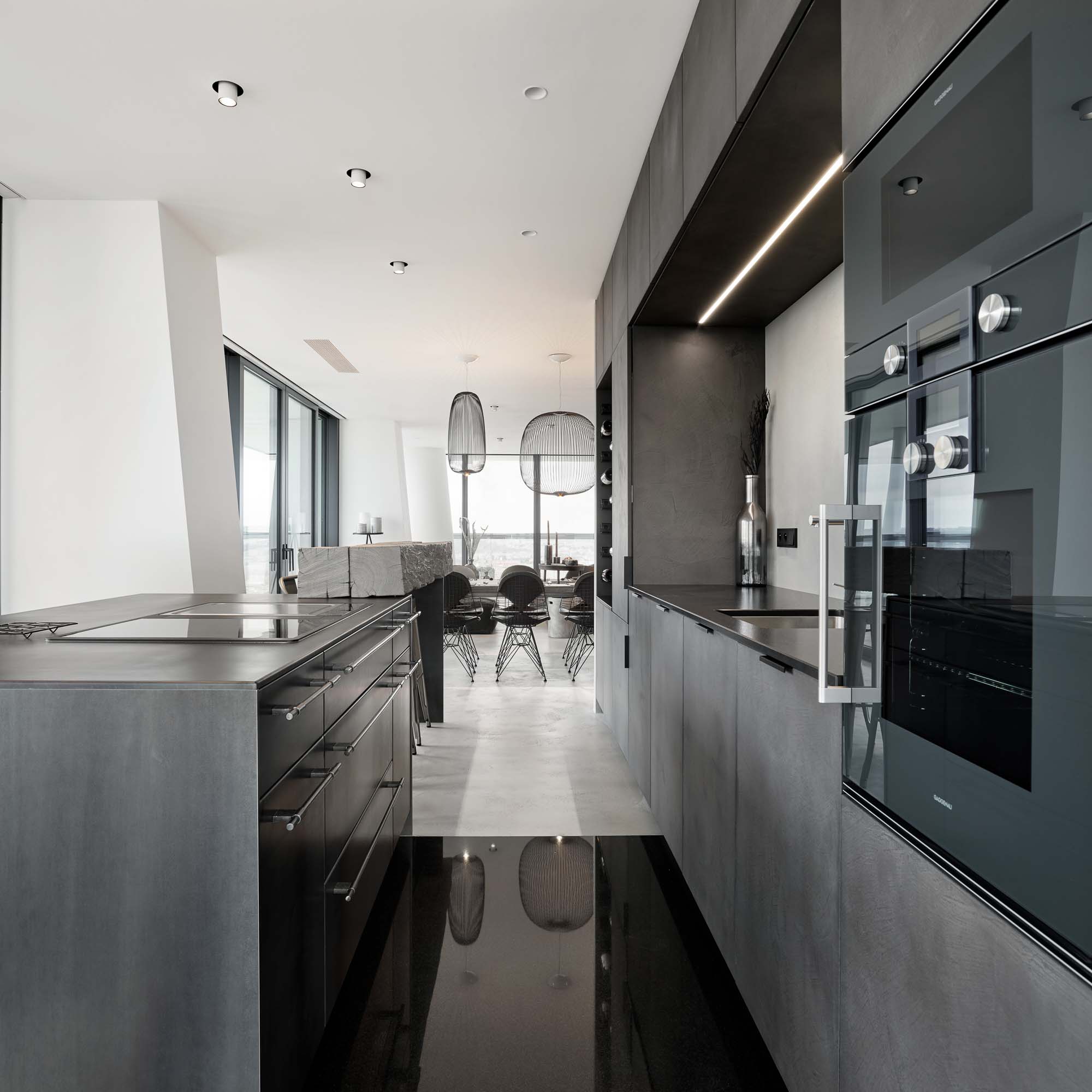 Through-view of the V-Tower Prague apartment kitchen – view across the kitchen island with Gaggenau appliances toward the dining and living areas, rattan pendant lights and panoramic windows