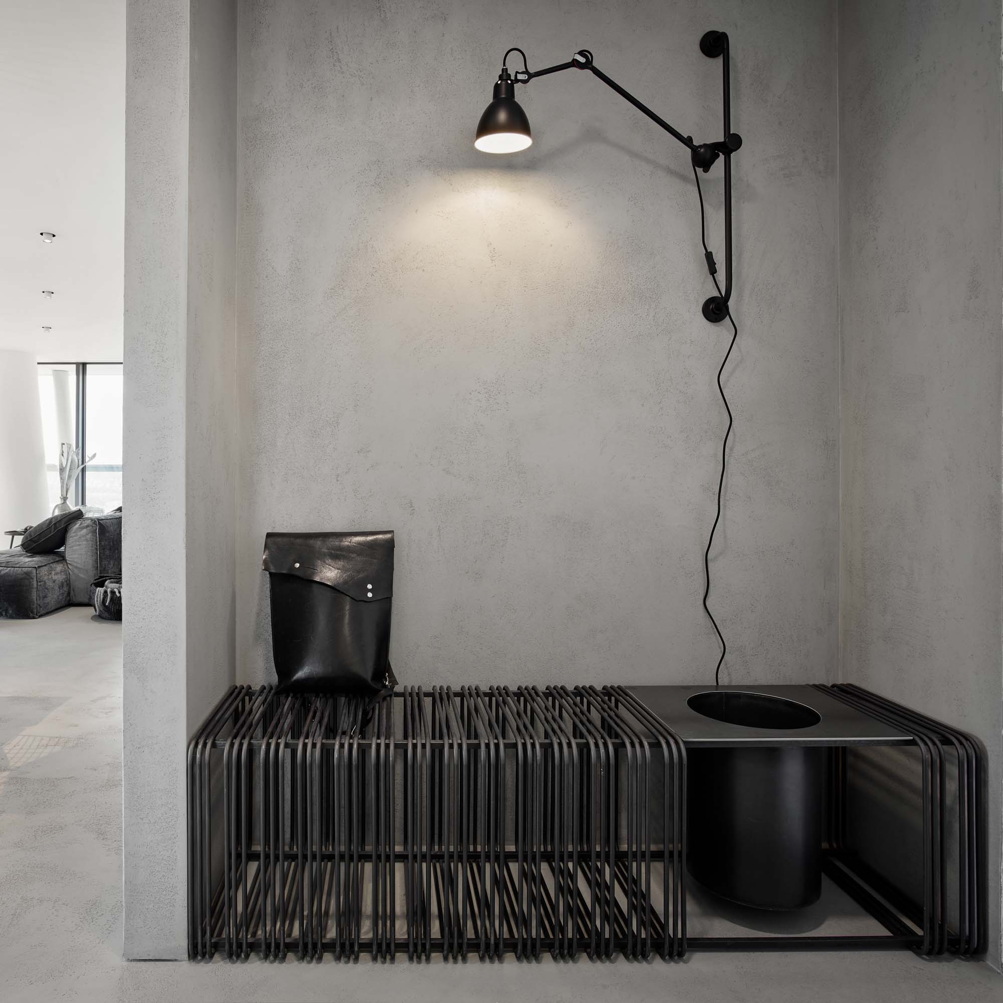 Entrance hallway of the V-Tower Prague apartment – black wire console table with leather bag, articulated wall lamp, grey concrete plaster on the wall and view through to the living room