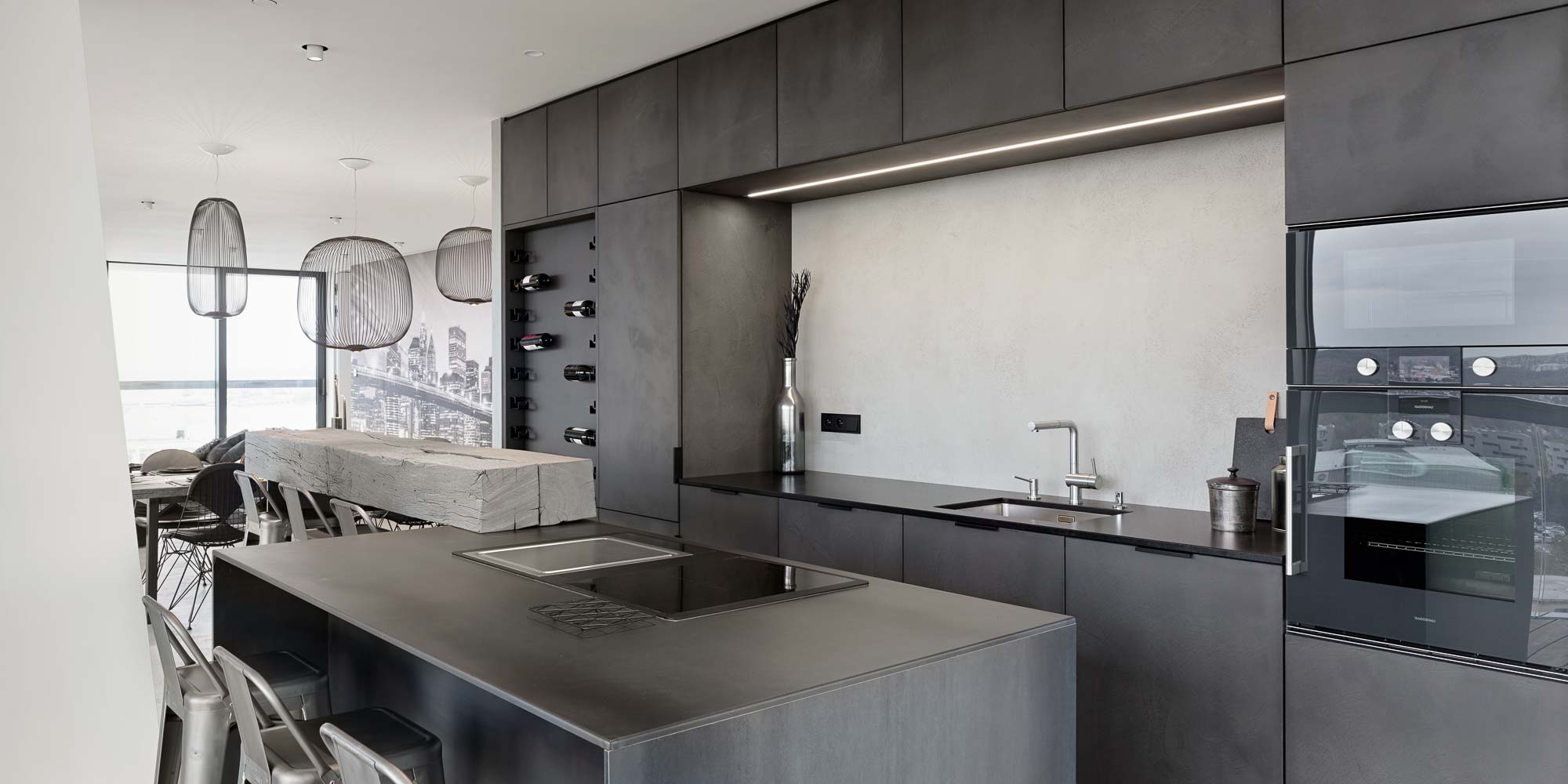 Kitchen and dining area of the V-Tower Prague apartment – overview of the dark kitchen with island, LED strip under cabinetry, wine rack niche, reclaimed wood bar, Gaggenau appliances, rattan chandeliers and NYC skyline mural in the background