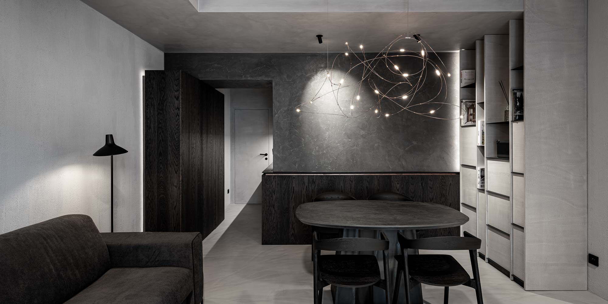 Celkový pohled na privátní lounge Selection Properties — tmavá pohovka, černý oválný stůl Ethnicraft Bok se čtyřmi černými dřevěnými křesly, lustr Moooi Flock of Light, vestavná knihovna vpravo