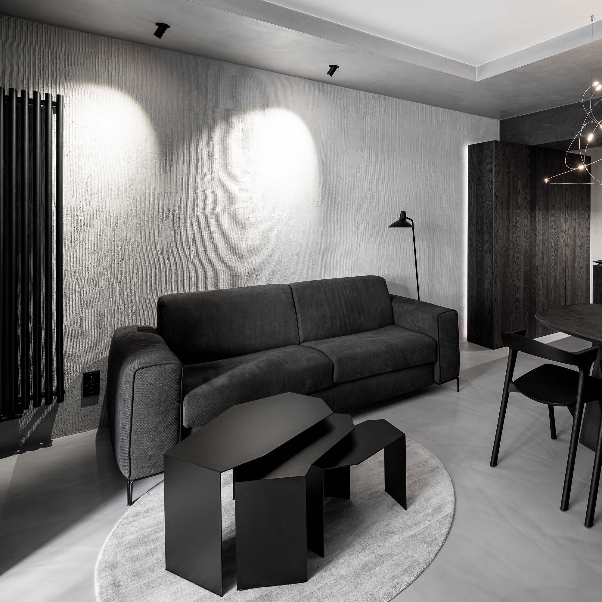 Lounge zóna Selection Properties — tmavě šedá rozkládací pohovka, asymetrická sada černých kovových odkládacích stolků, stojací lampička, kulatý koberec, černý vertikální radiátor vlevo, lustr Moooi Flock of Light